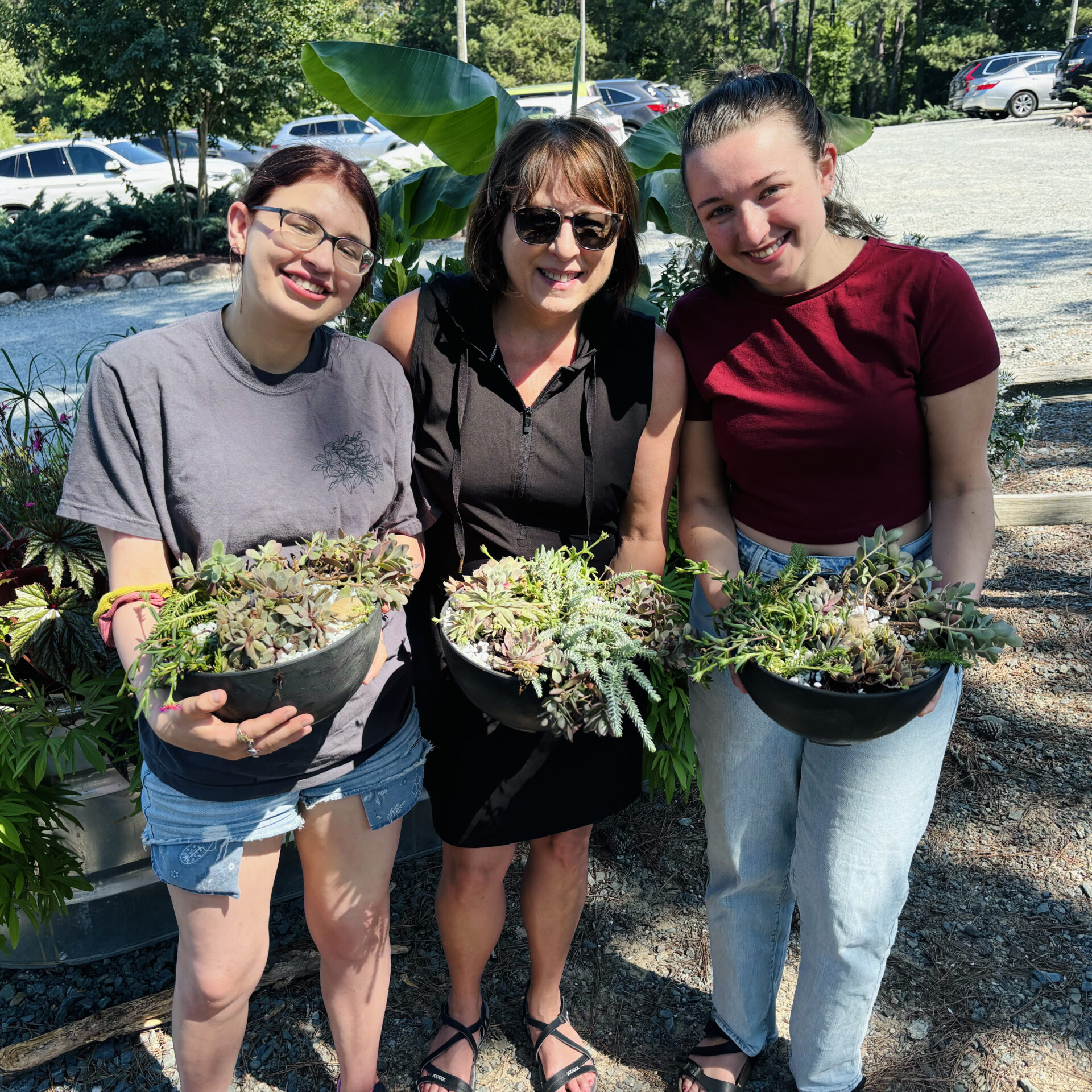 Three people outdoors holding succulent arrangements, surrounded by greenery and parked cars in a lot under bright sunlight. Casual, cheerful setting.