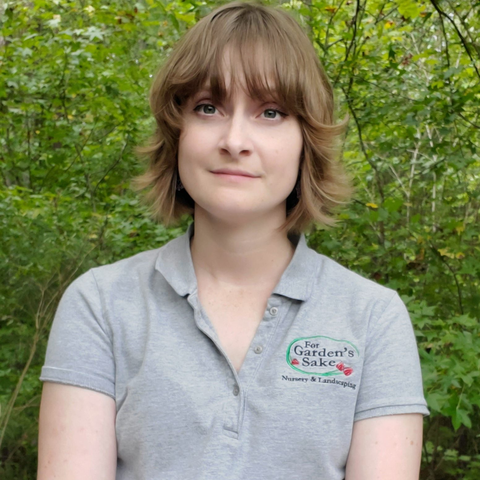 A person with short hair wears a gray polo shirt with a logo, standing in front of dense, green foliage.