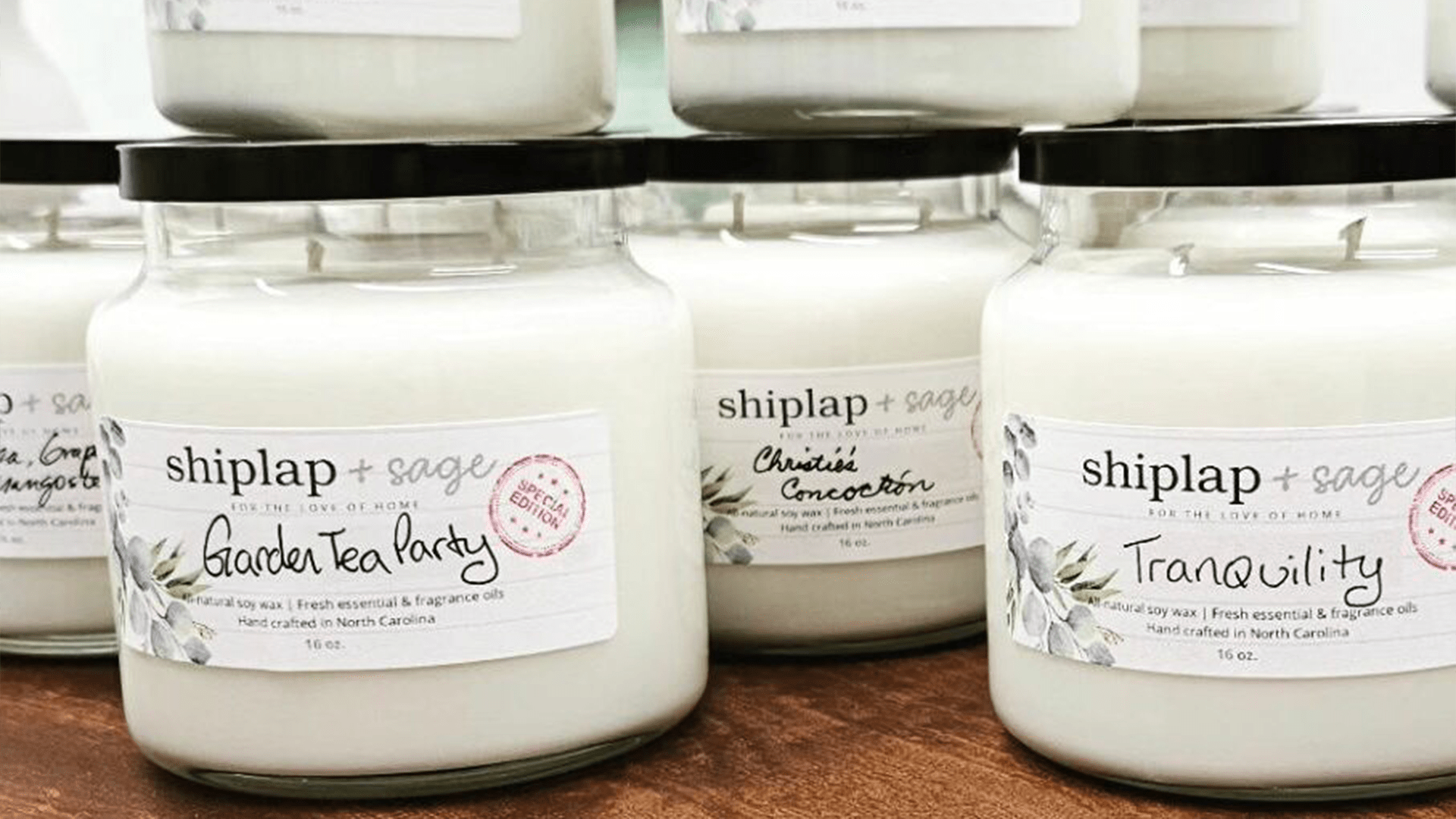 Rows of scented candles with labels featuring names like "Garden Tea Party" and "Tranquility." The jars have black lids and white wax.