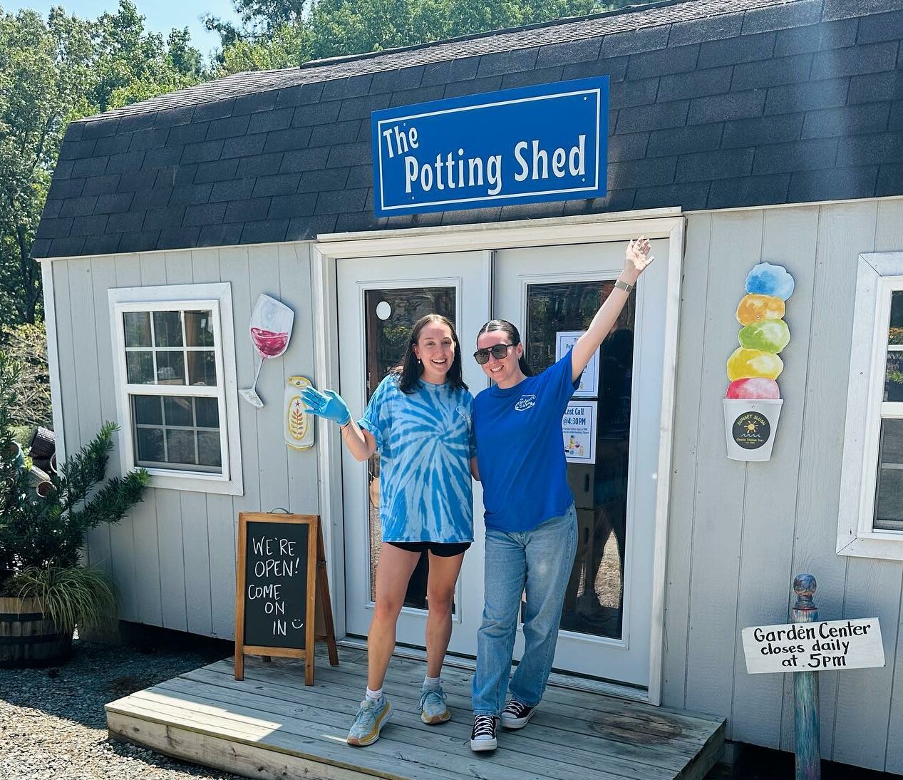 Two people smiling in front of "The Potting Shed," a small garden center. A sign reads, "We're Open! Come On In."