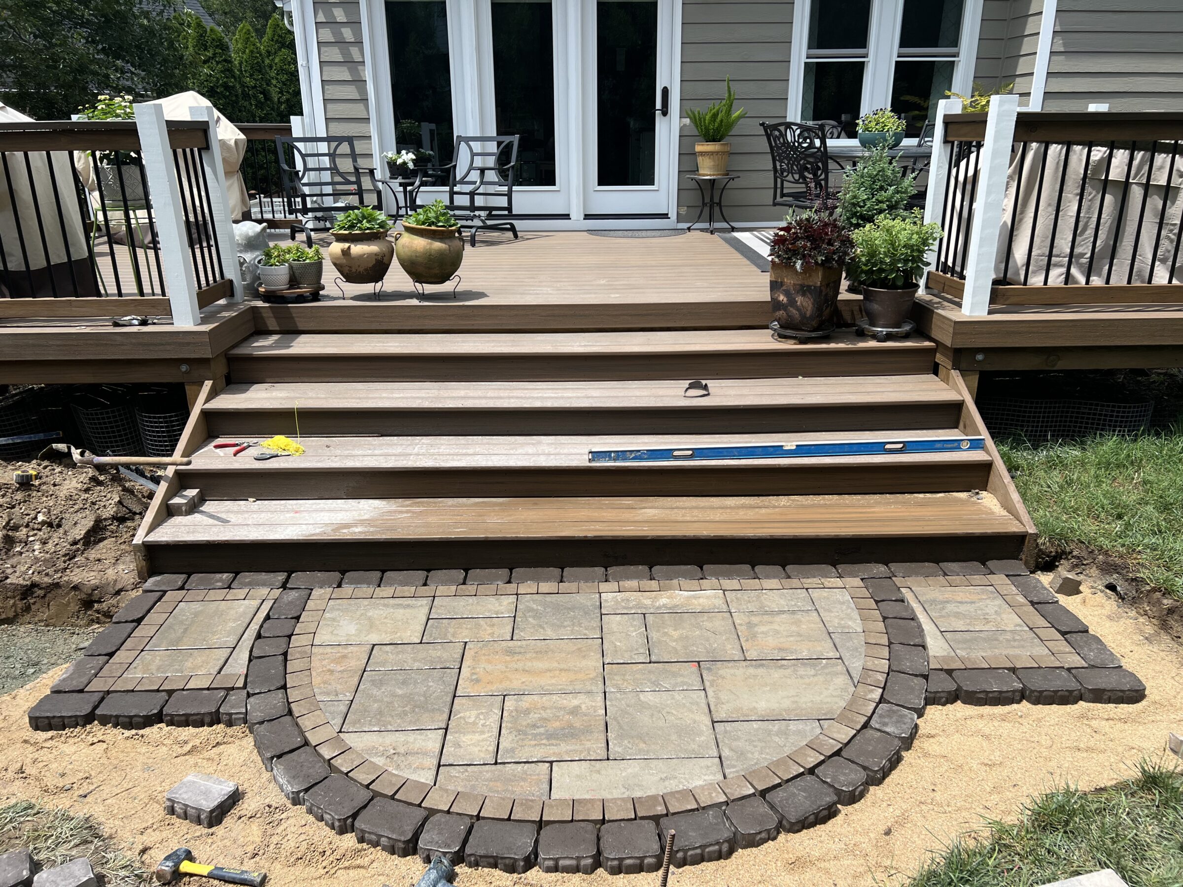 A wooden deck with potted plants leads to unfinished stone steps on sandy ground, featuring construction tools and surrounding greenery.