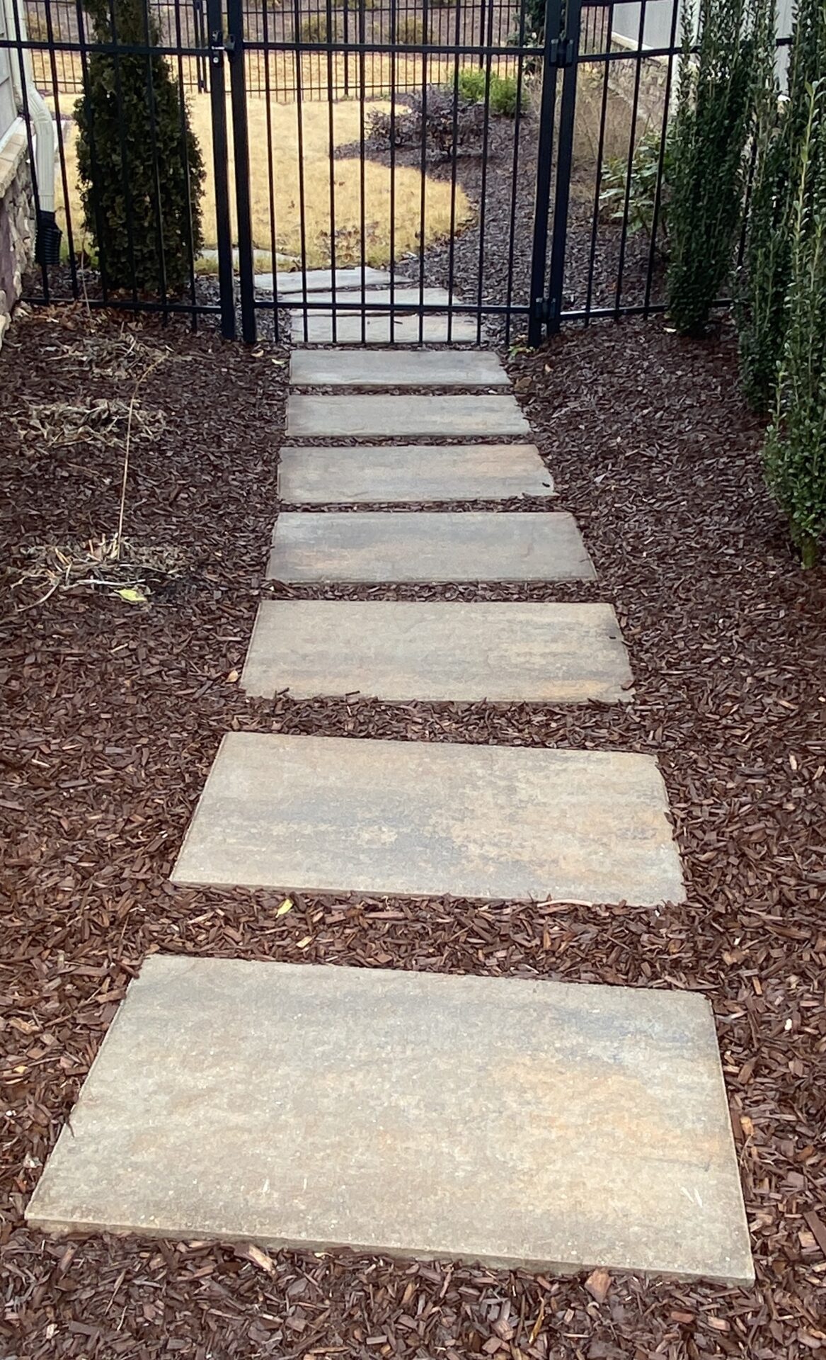 A stone pathway leads to a gated garden area, surrounded by mulch and greenery, with a yard visible beyond the gate.