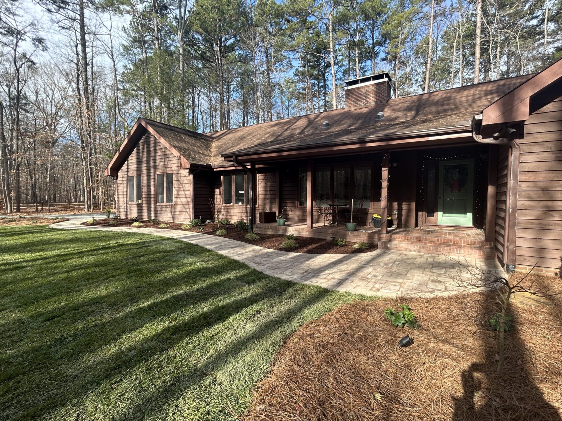 A rustic wooden house surrounded by trees, with a green lawn and brick pathway leading to the entrance under a bright sky.