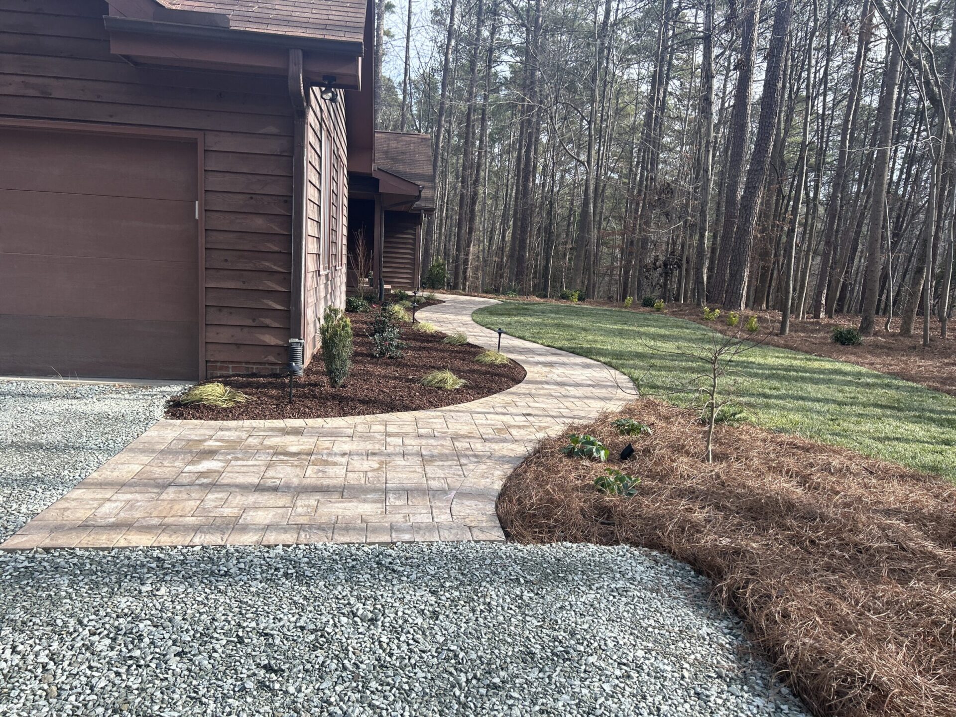 Wooden house with a stone pathway leading through landscaped garden beside a forest. The scene is tranquil with a rural atmosphere.