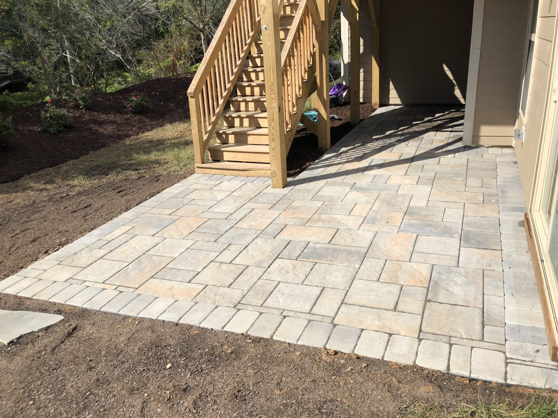 A sunlit outdoor area with a stone pattern patio and wooden staircase, surrounded by soil and greenery, under a clear sky.