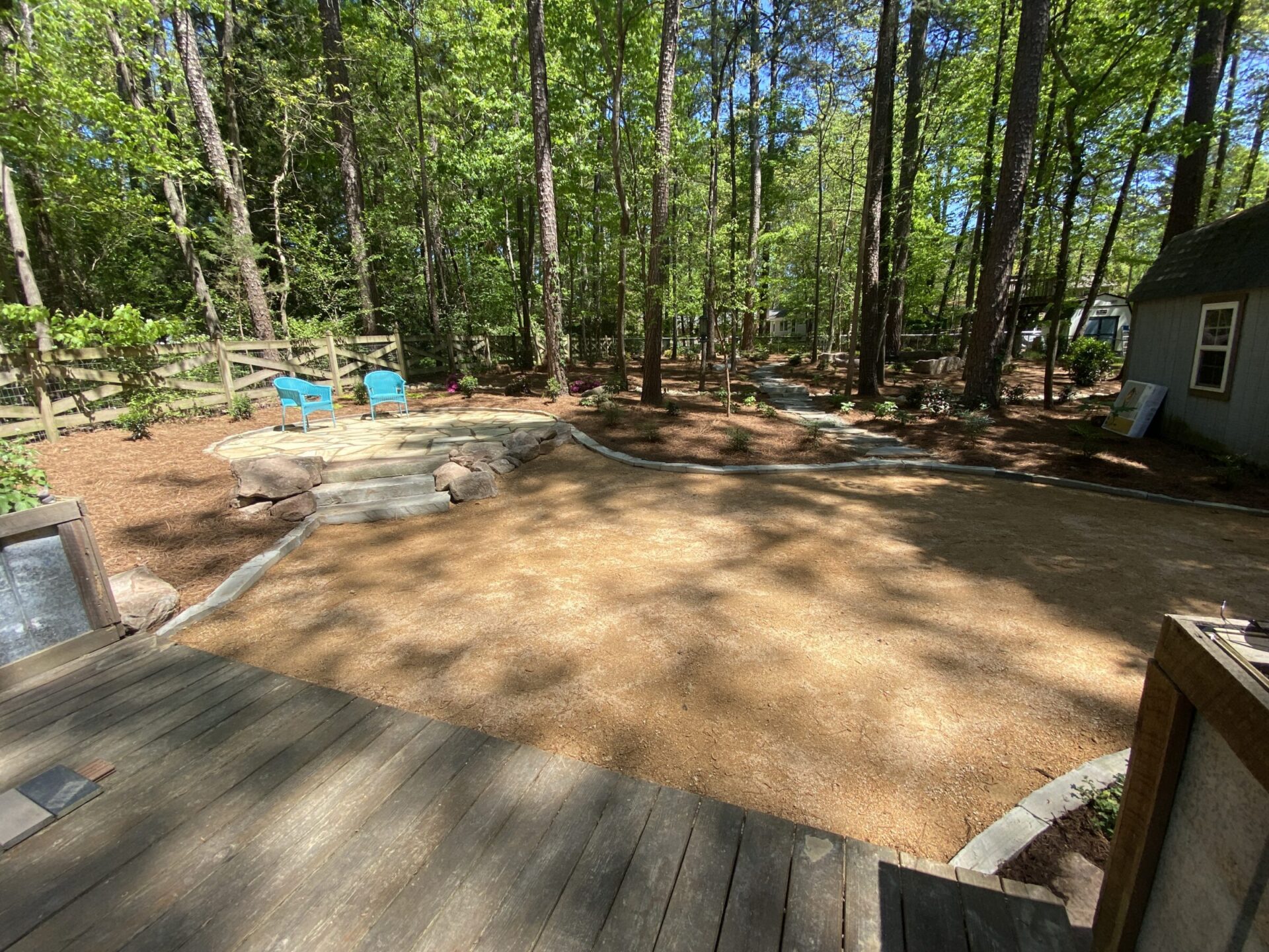 A wooded backyard features a gravel area, blue chairs, stone steps, and a small house, surrounded by tall trees and greenery.