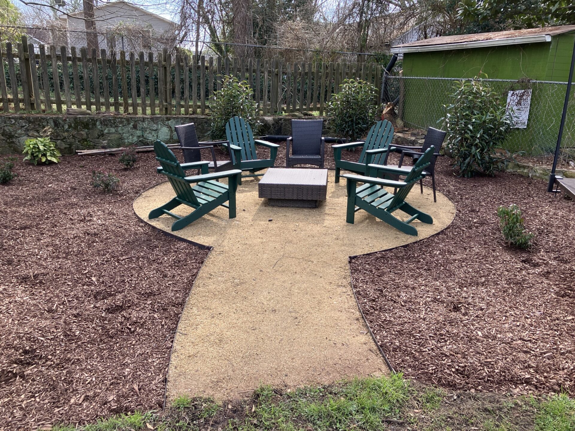 A backyard setting features green Adirondack chairs around a square fire pit, surrounded by mulch and a wooden and wire fence.