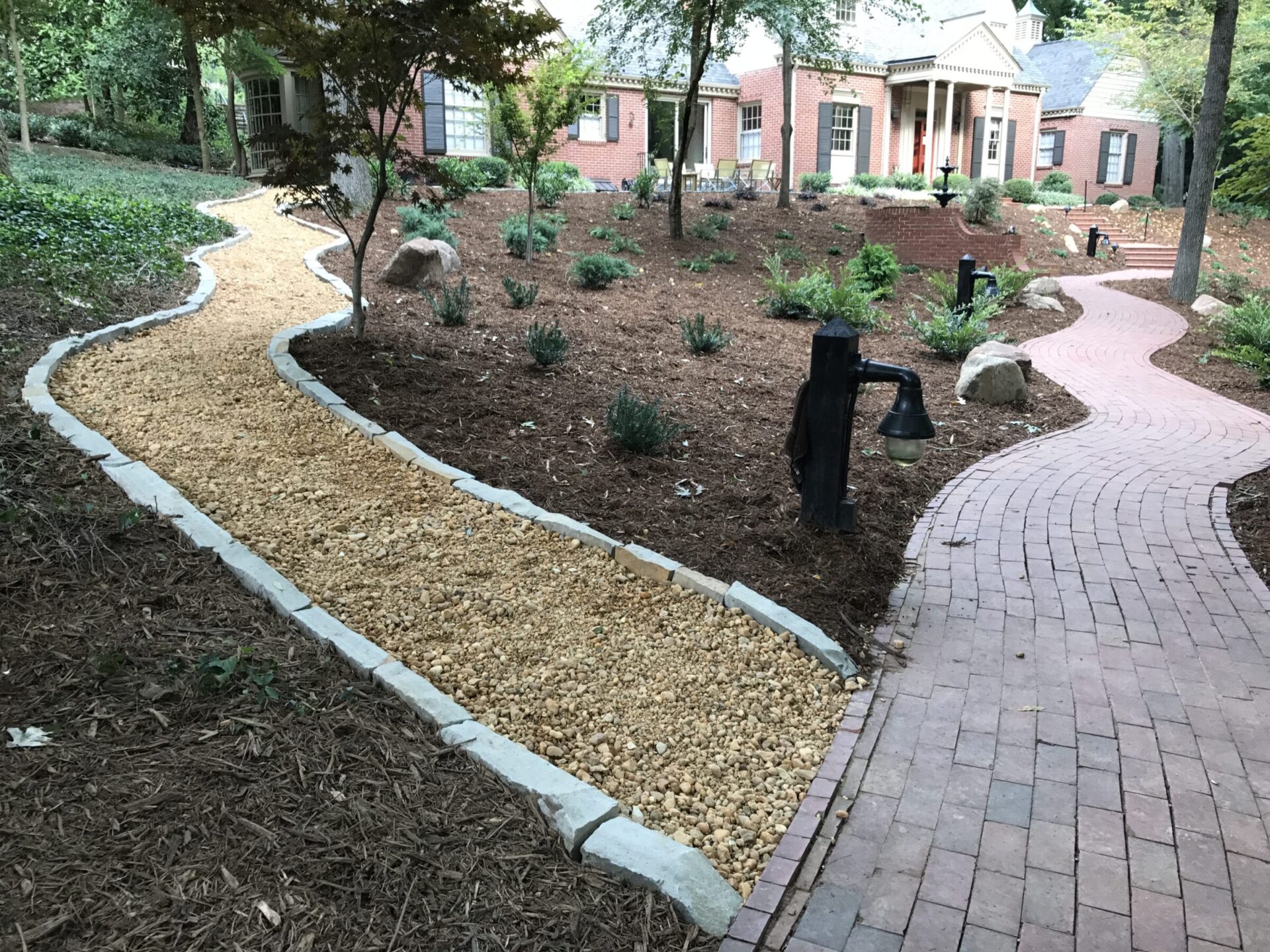 Curved gravel and brick paths lead to a brick house with columns, surrounded by landscaped gardens and trees, featuring a vintage-style lamp.