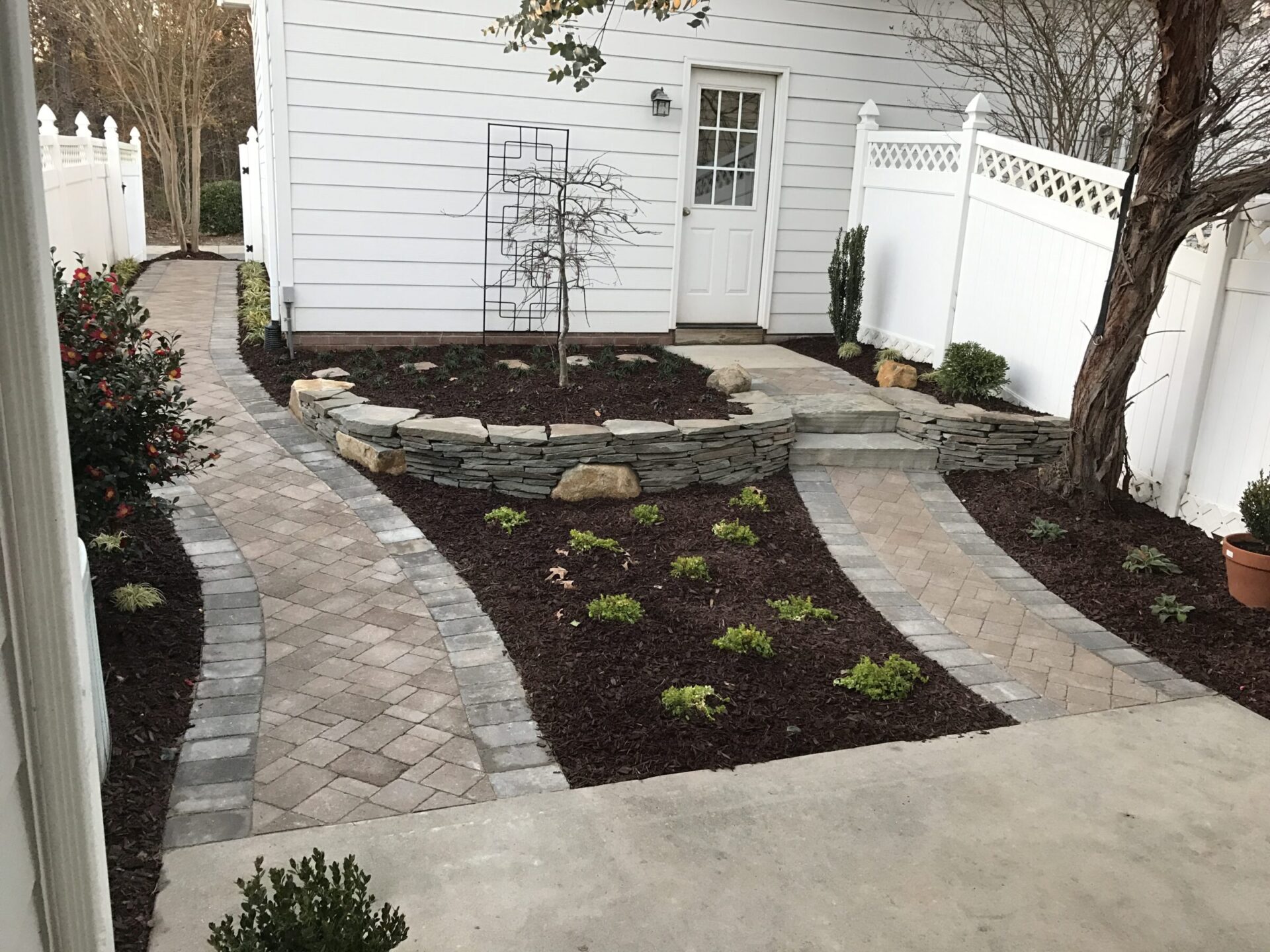 A neat backyard with paved paths, white fencing, and a small garden featuring shrubs and a central rock installation near a white building.