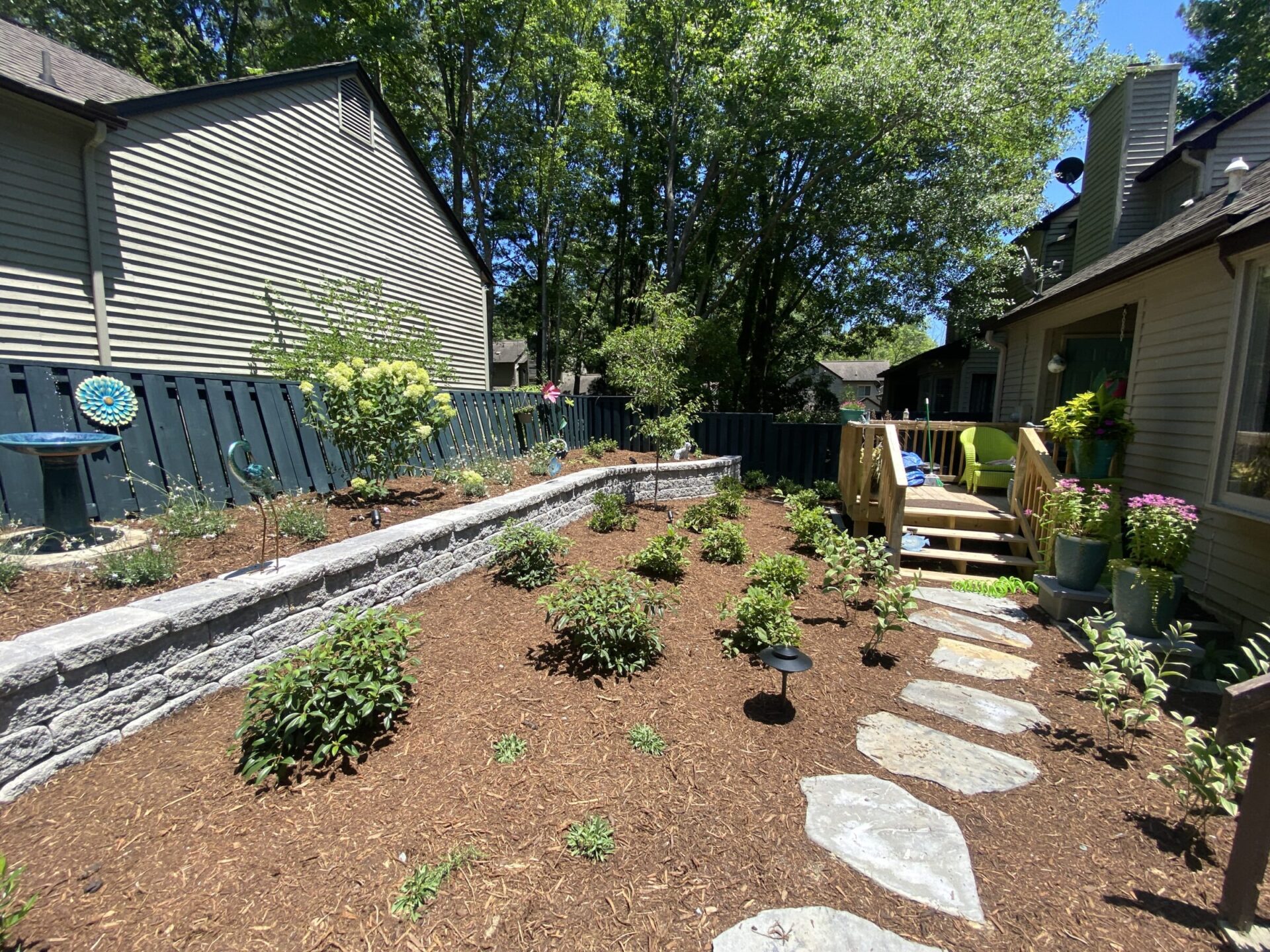 A backyard garden features a stone path, newly planted shrubs, wooden steps, and fences, surrounded by two houses with green trees in the background.