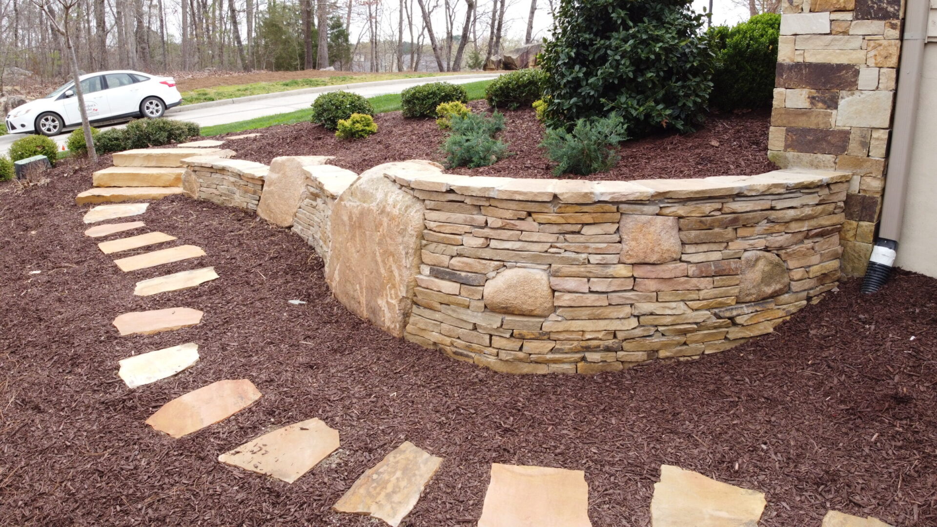 Stone wall with curved design and adjacent pathway made of stone slabs. Surrounded by mulch and shrubs, car parked in background.