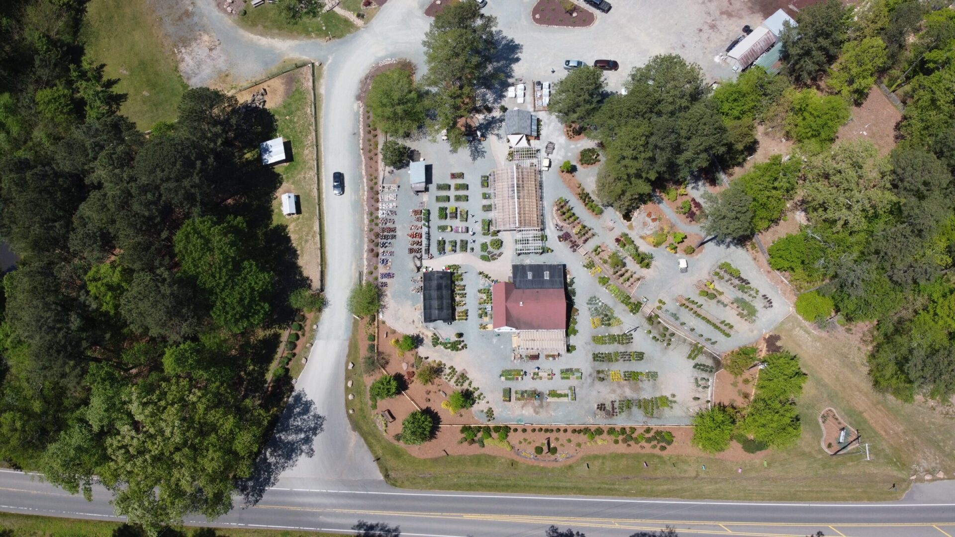 Aerial view of a garden center surrounded by trees with arranged plants, pathways, and a red-roofed building, adjacent to a curving road.