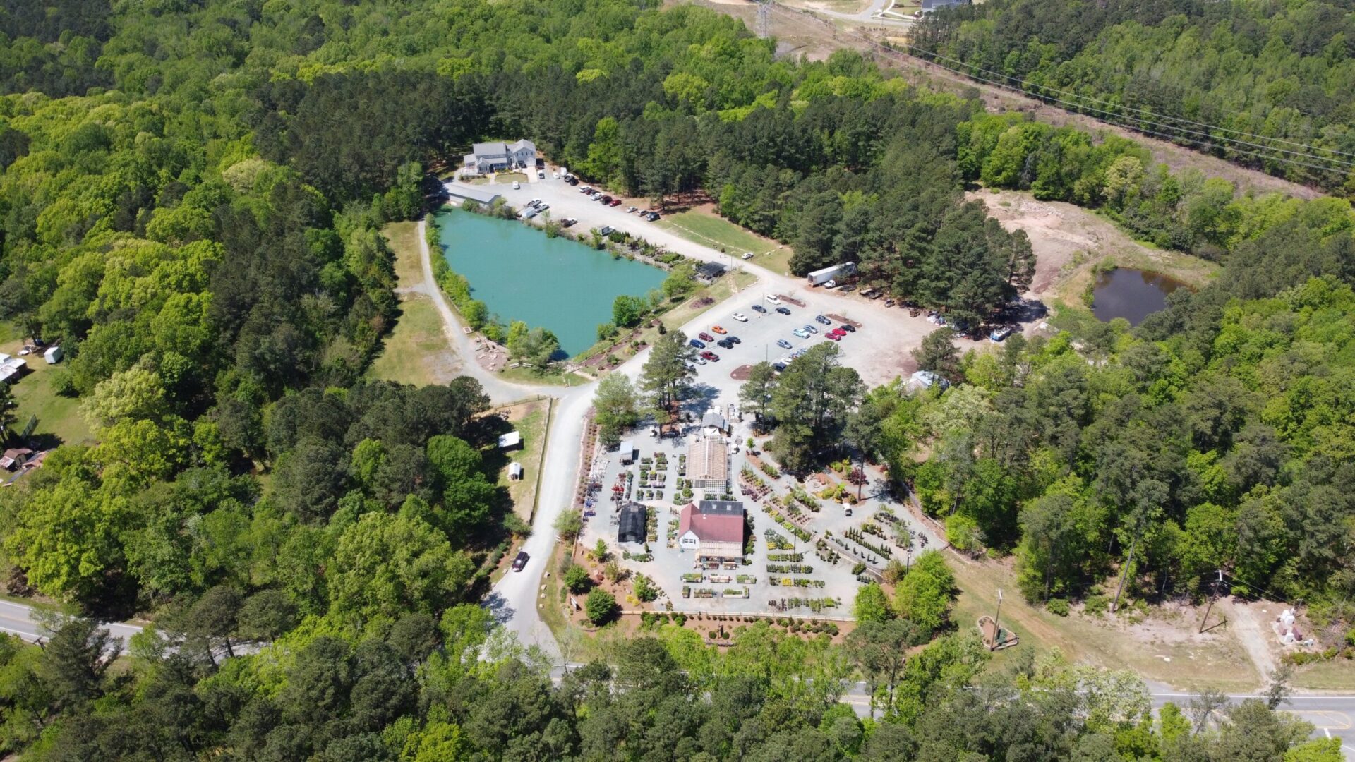 Aerial view of a lush green area with a landscaped garden, a pond, parking lot, and several buildings surrounded by dense forest.