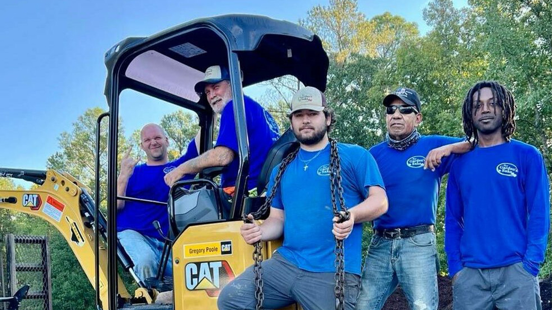 Four people in blue shirts stand and sit on a yellow construction vehicle, outdoors with trees in the background.