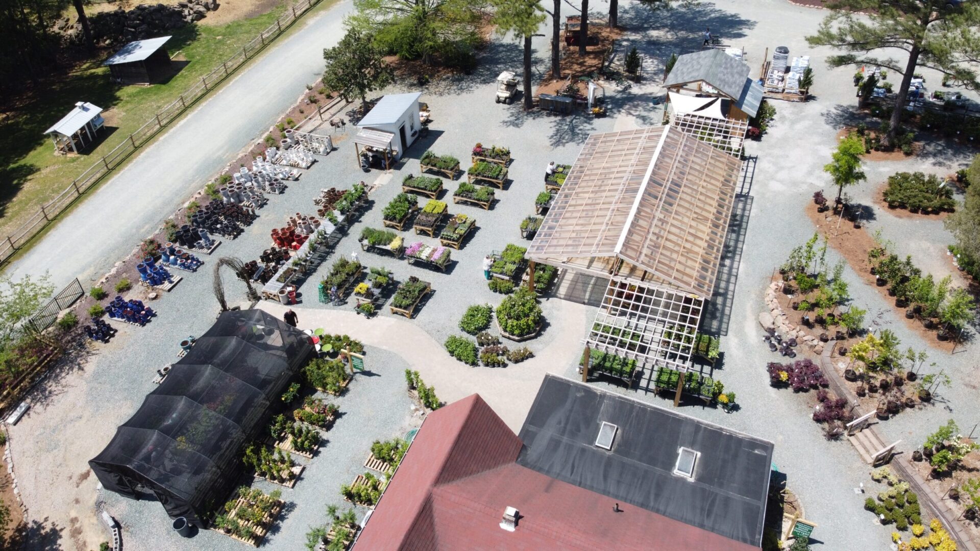 Aerial view of a nursery featuring greenhouses, various plant sections, and pathways. Surrounded by trees and bordered by a gravel road.