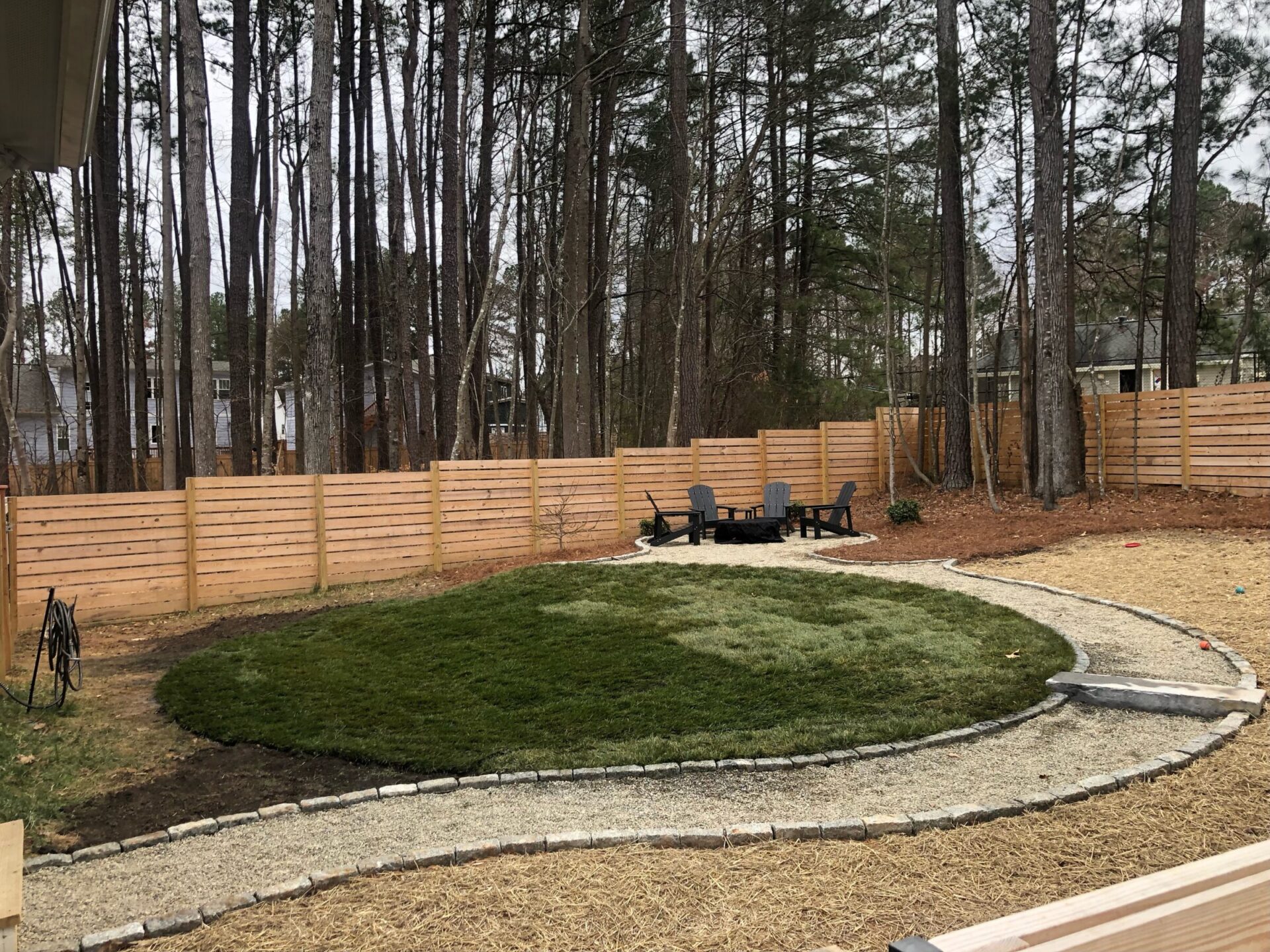 A landscaped backyard with circular grass, surrounded by a gravel path, trees, wooden fence, and Adirondack chairs around a small fire pit.