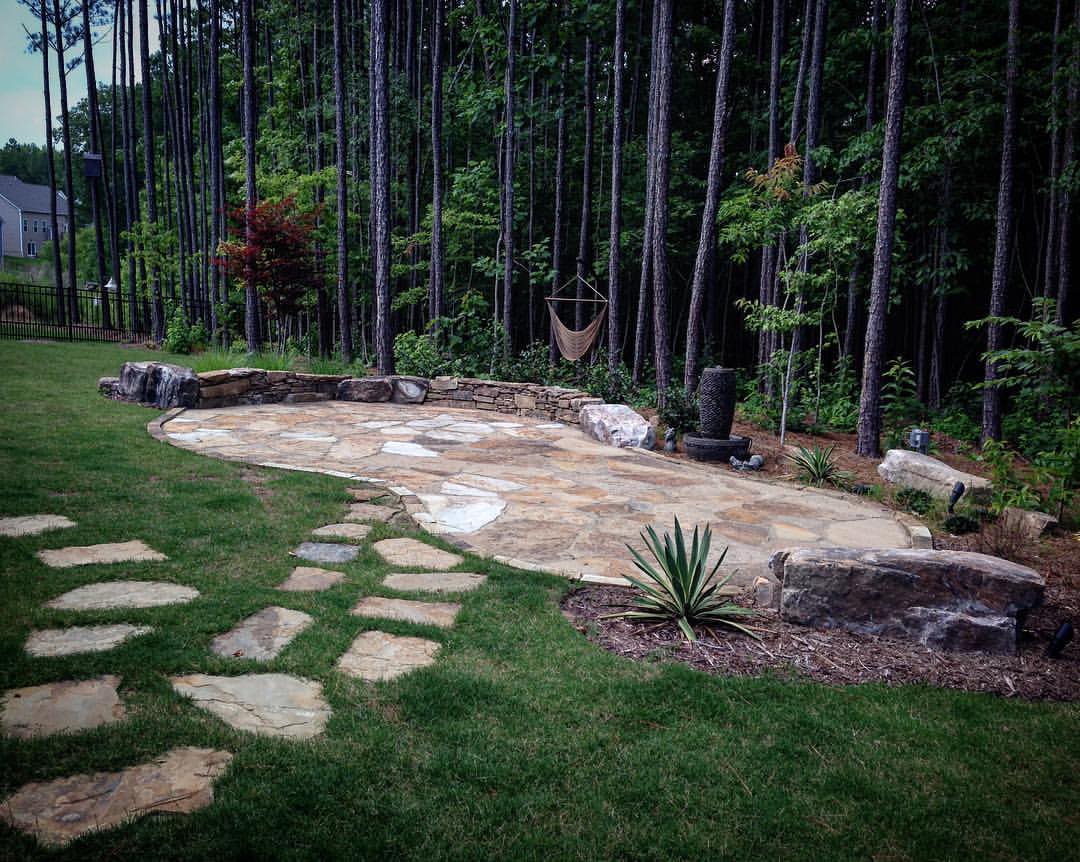 Stone patio in a backyard surrounded by tall trees and greenery. Rock path leads through landscaped garden with small plants and a cozy hammock.