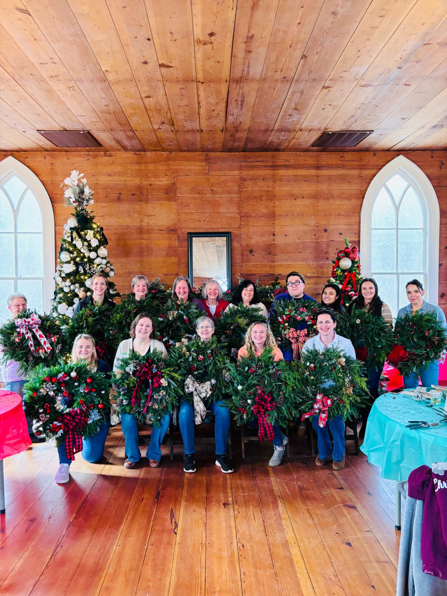 A group of people holding Christmas wreaths in a wooden room with a decorated tree and arched windows, celebrating the holiday season.