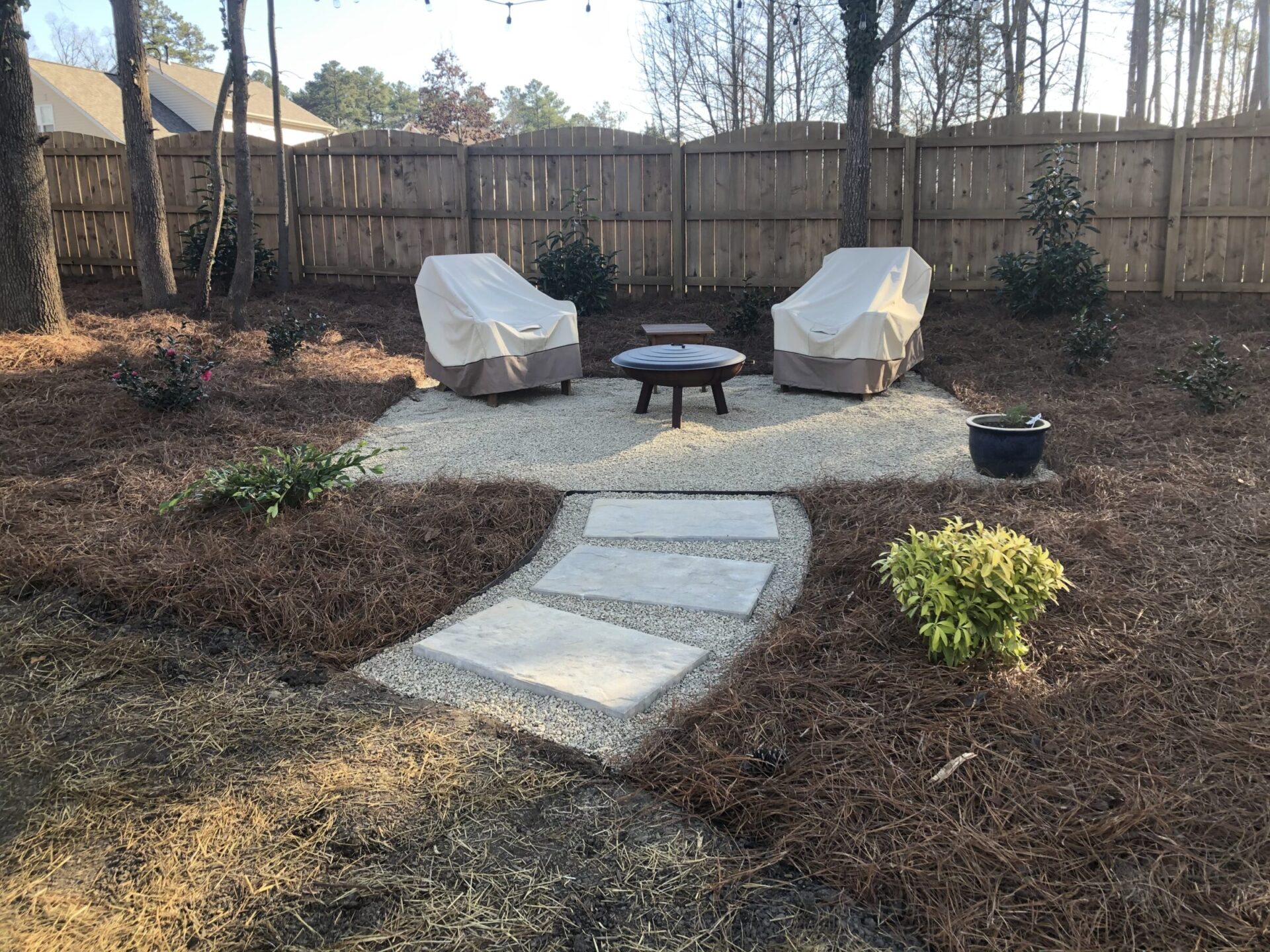 Cozy garden setting with two covered chairs, fire pit, and pathway. Surrounded by trees, mulch, and plants, enclosed by a wooden fence.