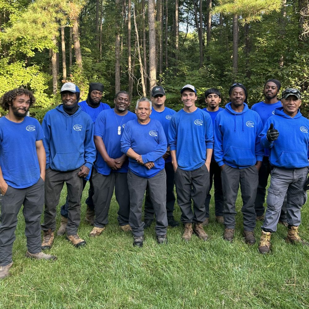 A group of ten people wearing blue shirts and caps stand on grass, in front of a forested area with tall trees.