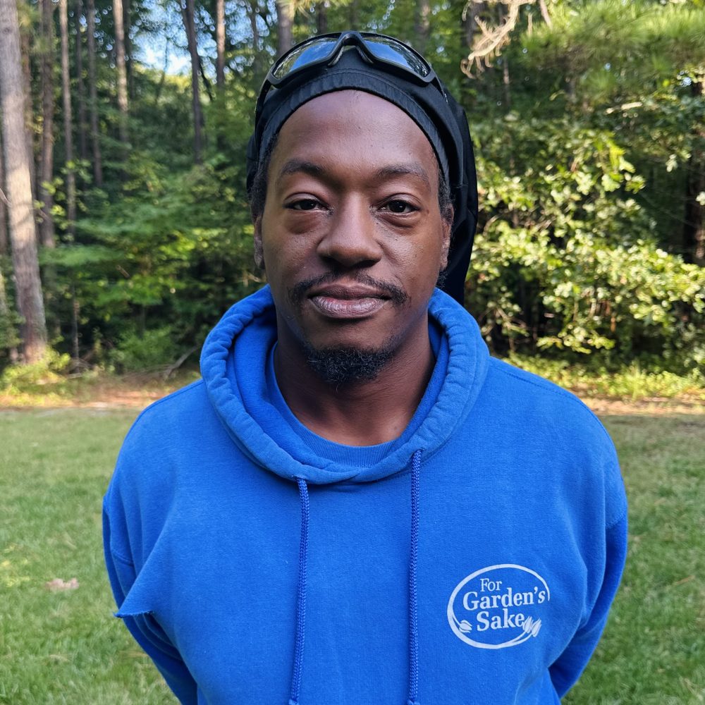 A person in a blue hoodie stands outdoors, surrounded by lush trees and grass, wearing sunglasses on their head, smiling at the camera.