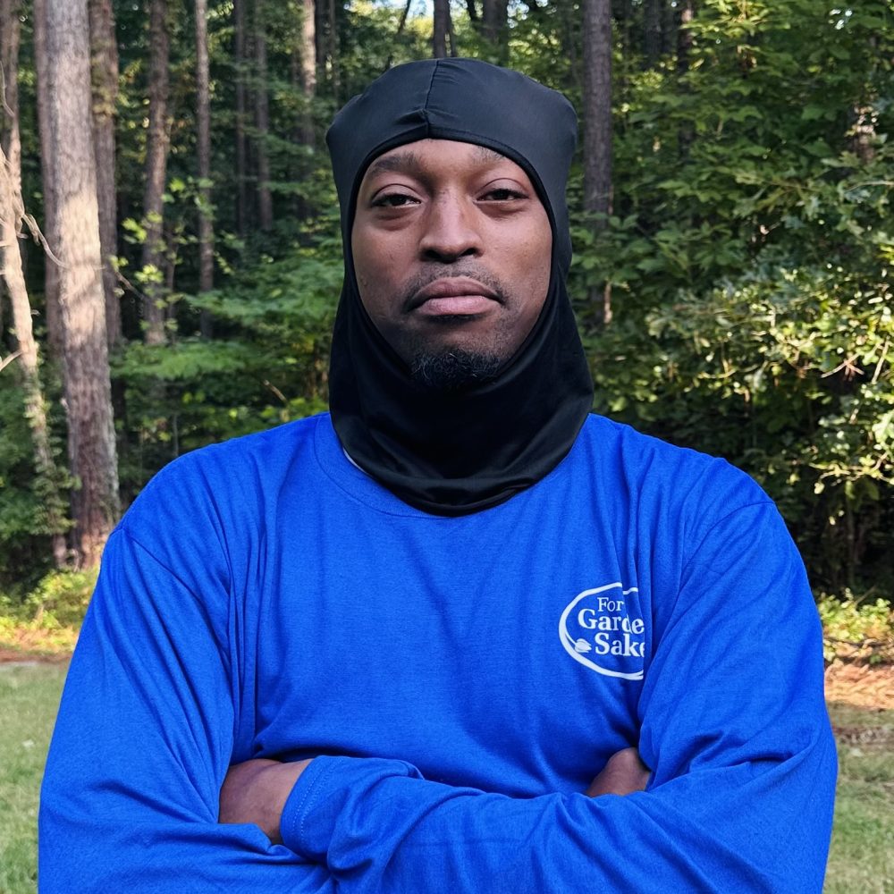 A person in a blue shirt stands in a wooded area, with trees in the background, looking directly at the camera.