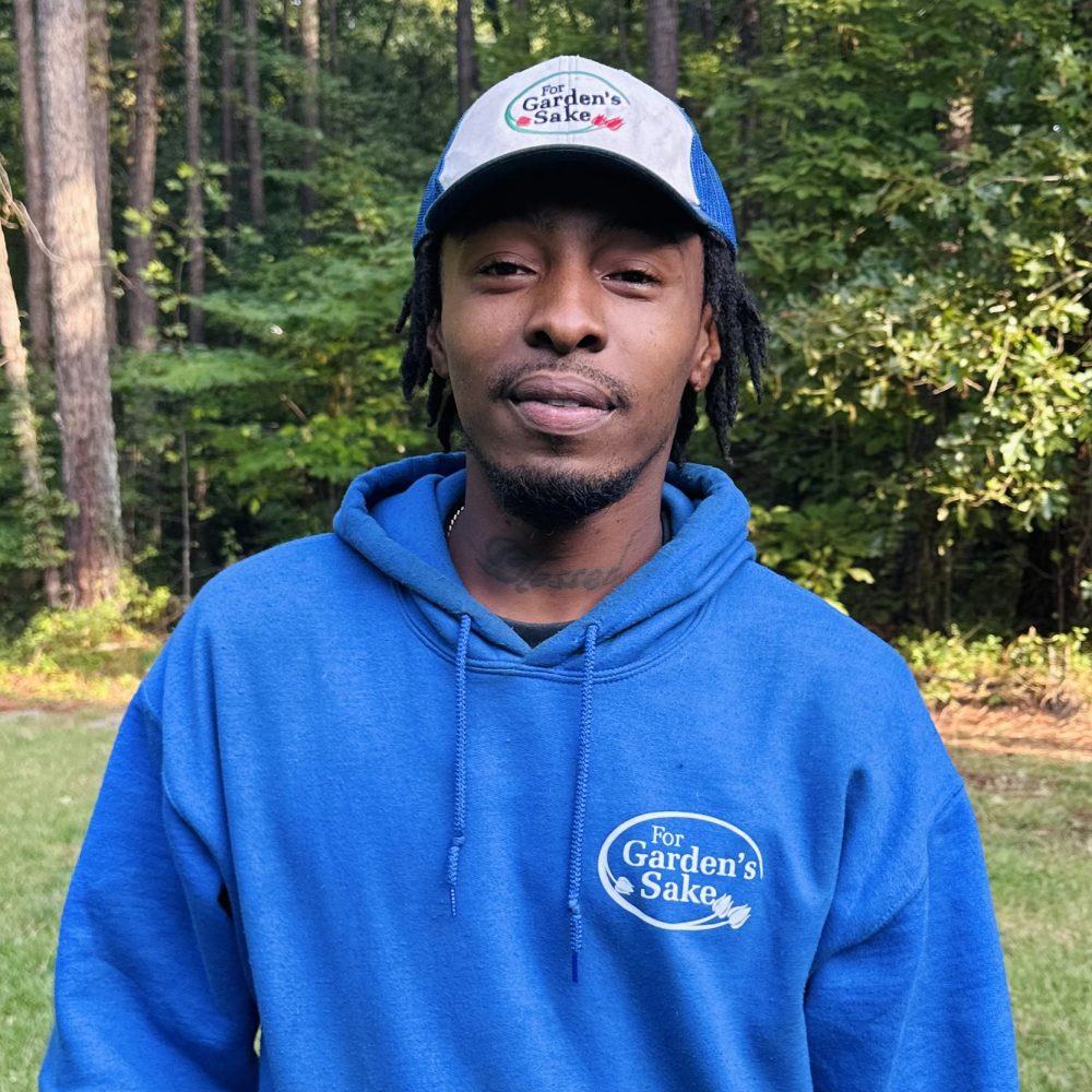 A person wearing a blue "For Garden’s Sake" hoodie and cap stands outdoors, surrounded by trees and greenery.