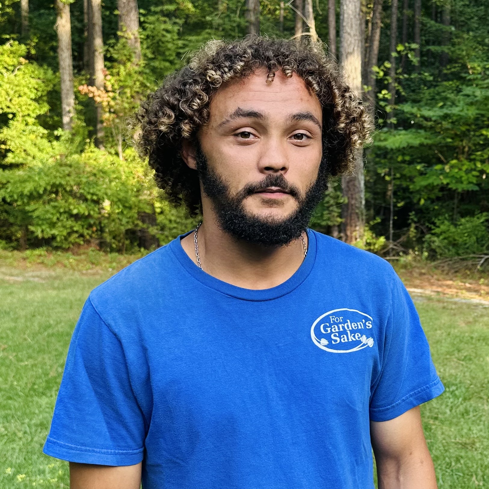 A person with curly hair and a beard stands in a grassy area, wearing a blue shirt with text. Trees are visible in the background.