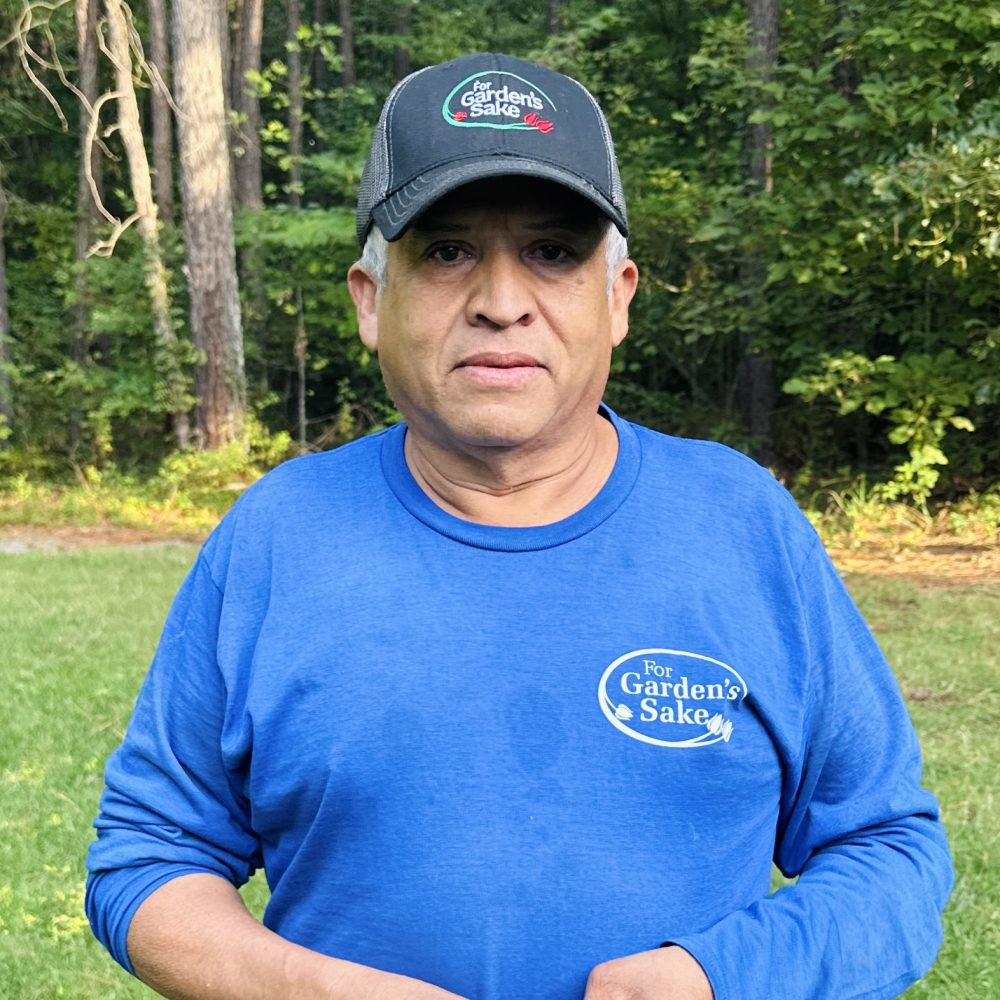 A person wearing a blue "For Garden's Sake" shirt and cap stands outdoors, surrounded by trees and grass in a forest setting.