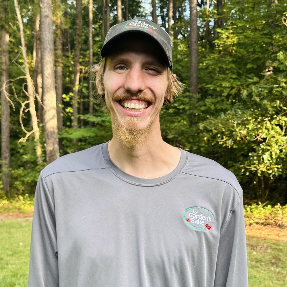 A person wearing a logo-branded shirt and cap smiles in a wooded area. Sunlight filters through the trees in the background.