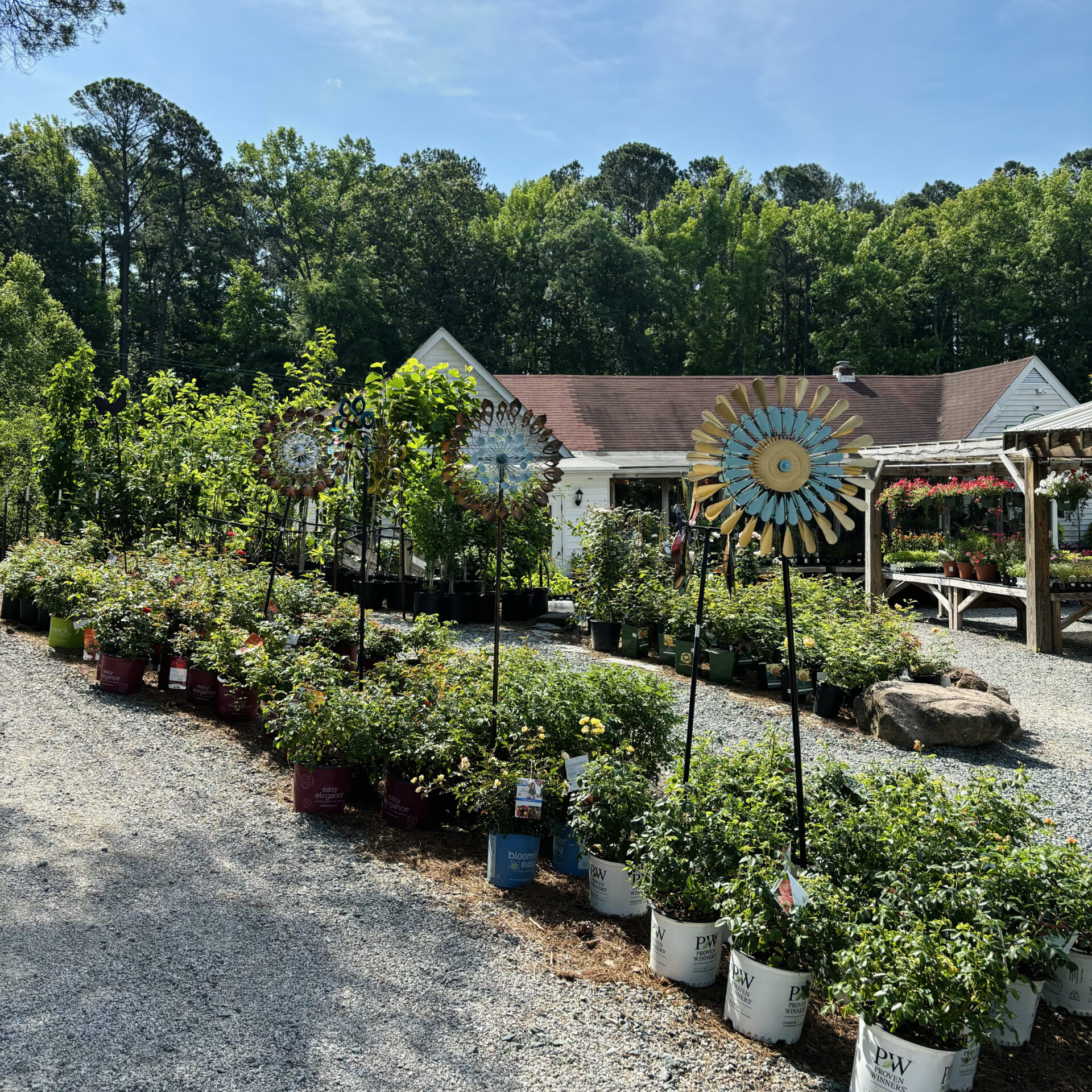 Outdoor garden center with colorful potted plants, metal wind spinners, and a rustic building. Surrounded by lush trees and gravel paths.