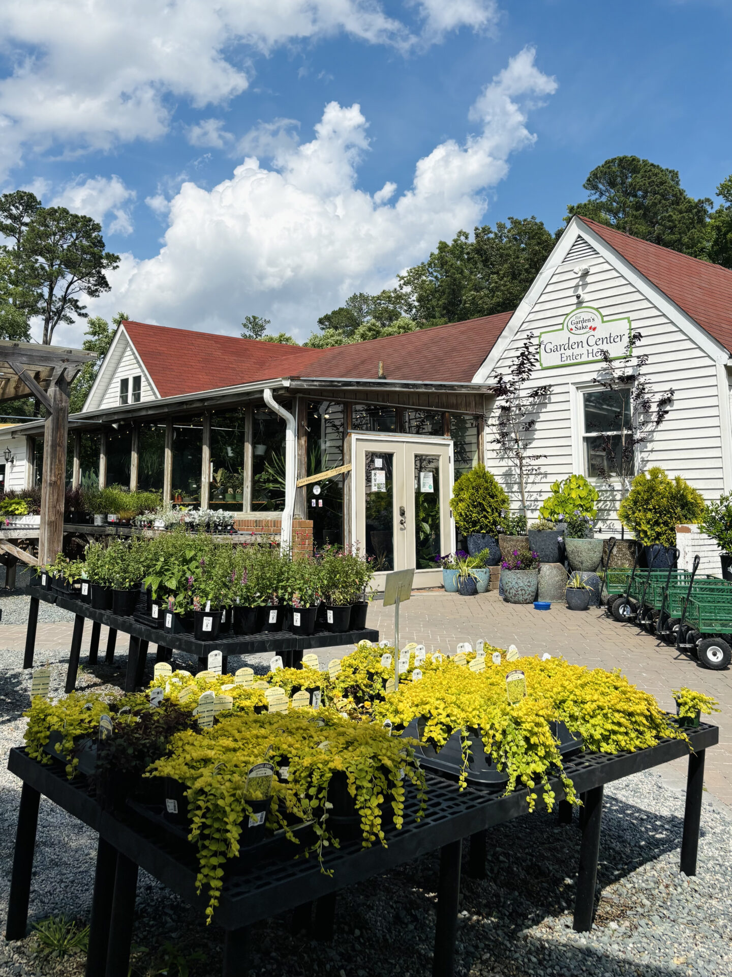 A garden center with a rustic building, red roof, and outdoor plants under a blue sky with clouds. Potted plants and carts visible.