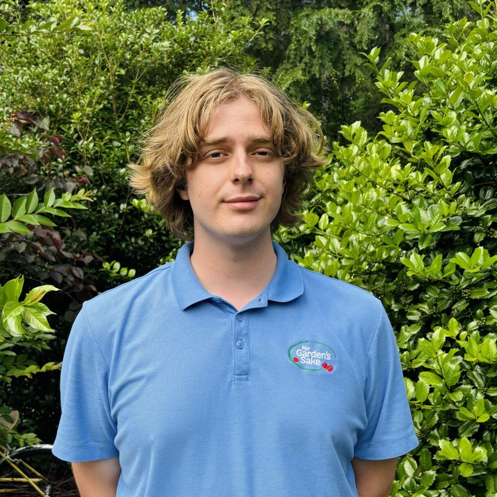 A person in a blue polo shirt stands in front of lush green foliage, with a calm expression and natural outdoor setting.