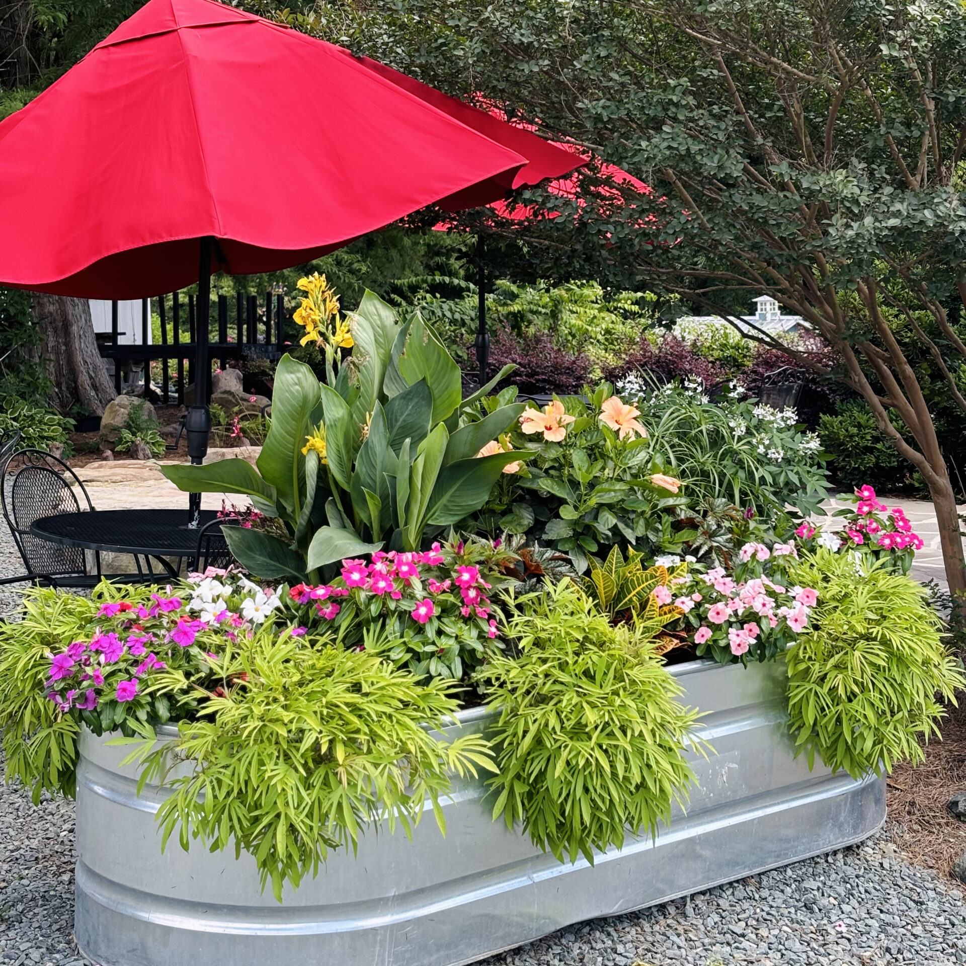 Lush garden with vibrant flowers in a metal planter, beneath a red umbrella. Patio furniture and trees create a serene outdoor space.