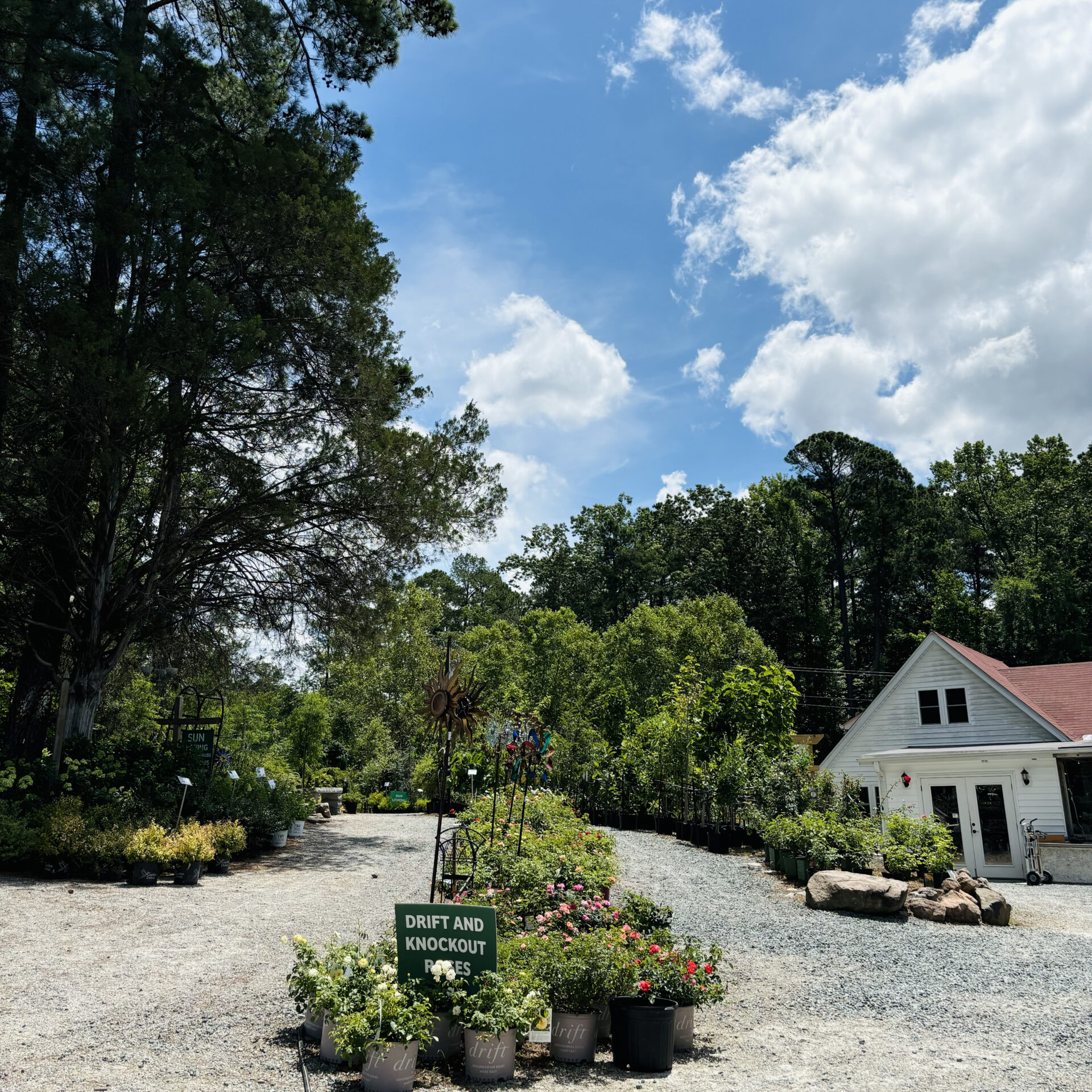 A garden center with gravel path, flower pots, and a small white building surrounded by trees on a clear, sunny day.