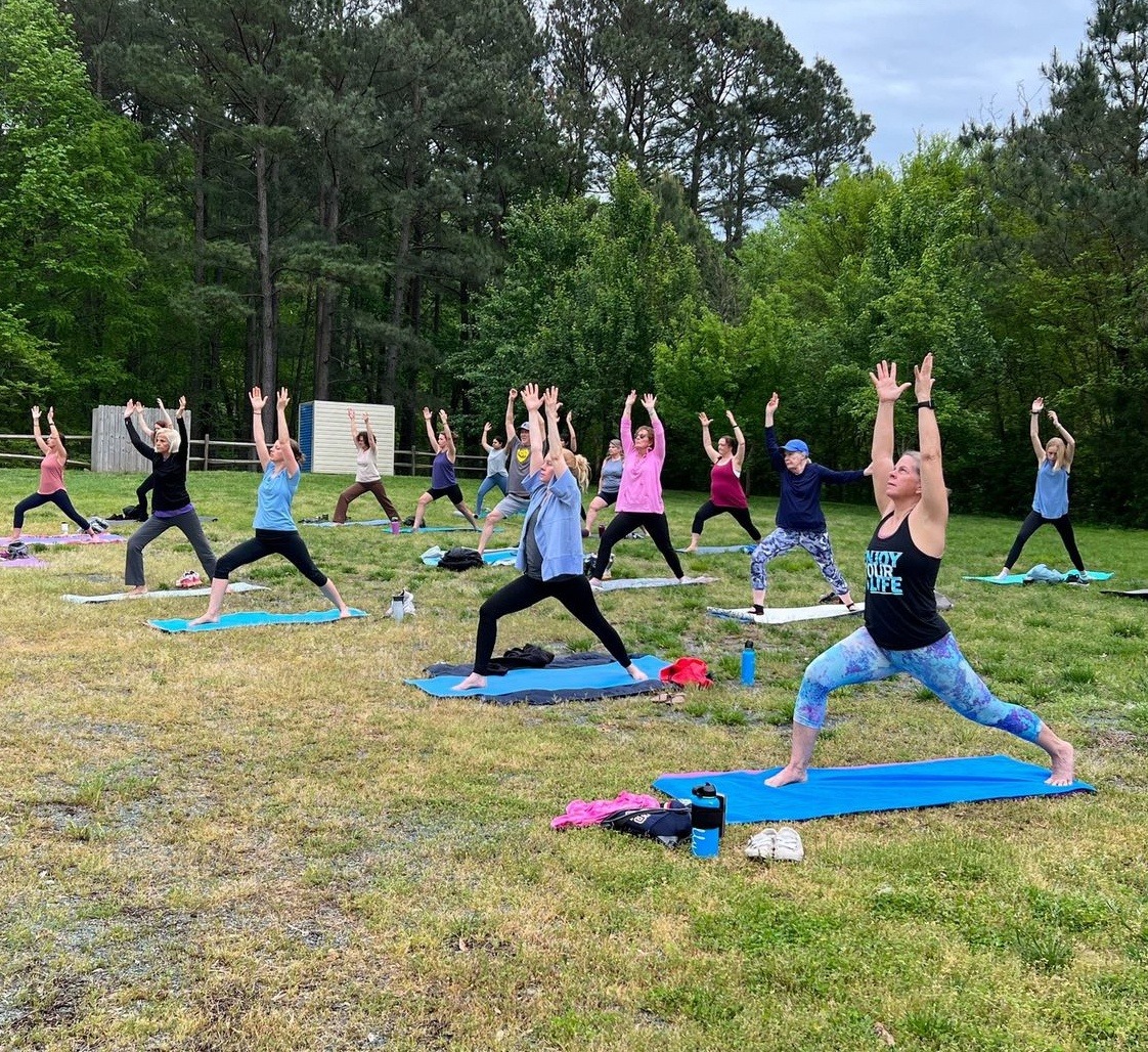 A group of people practices yoga outdoors on mats in a grassy field surrounded by trees, under a cloudy sky.