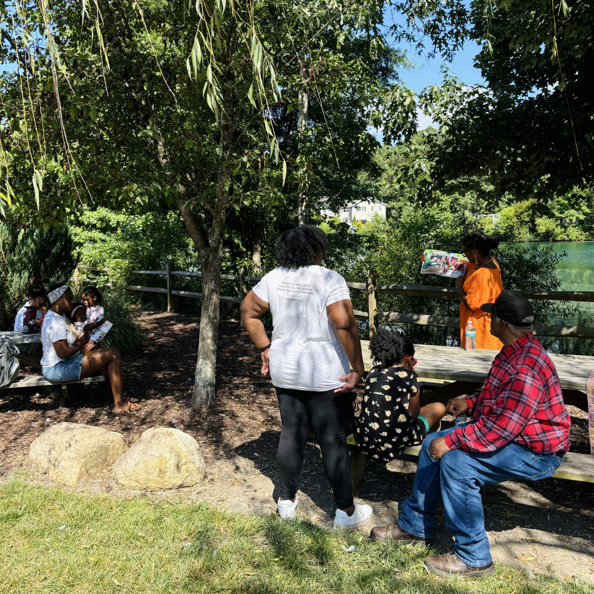 A group of people gather under trees in a park, listening to a story being read aloud. Picnic table and rocks nearby.