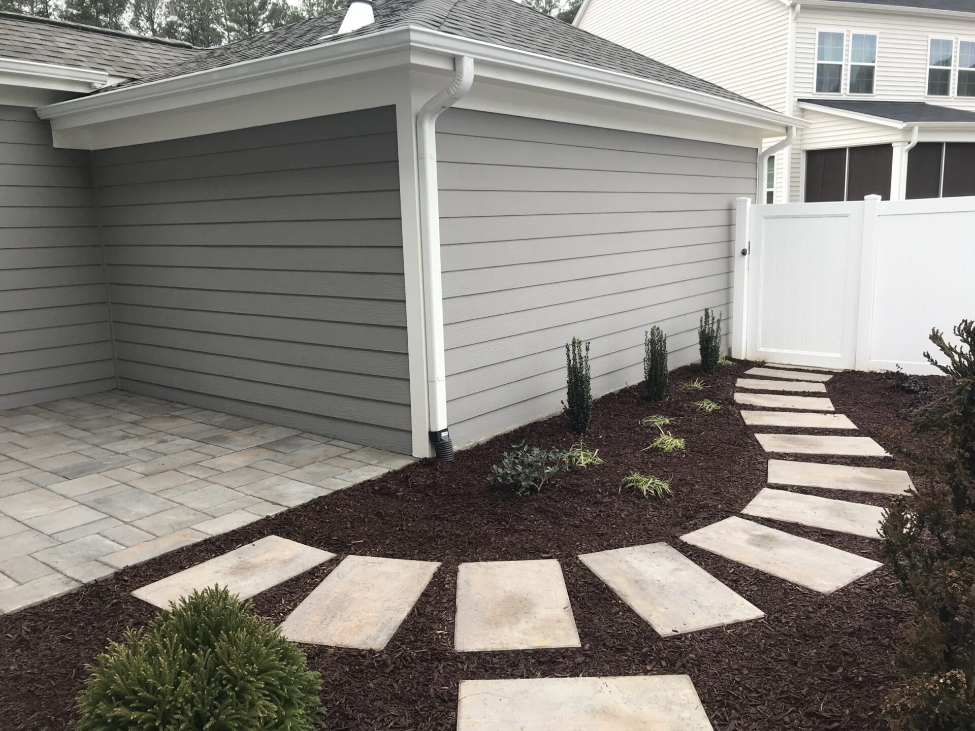 A modern backyard features a paved stone path, landscaped garden, gray siding, and a white fence, creating a clean and organized outdoor space.