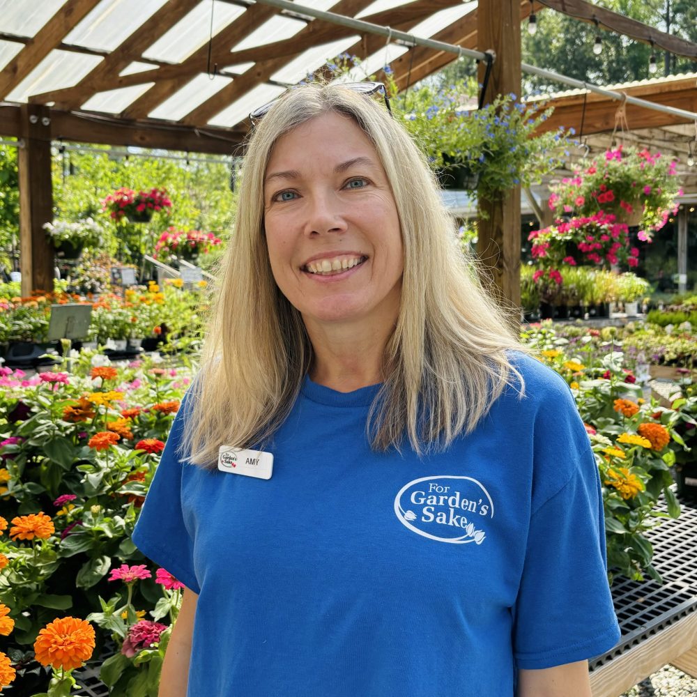 A person wearing a blue shirt stands smiling in a garden center surrounded by vibrant flowers and plants. Wooden structures overhead.