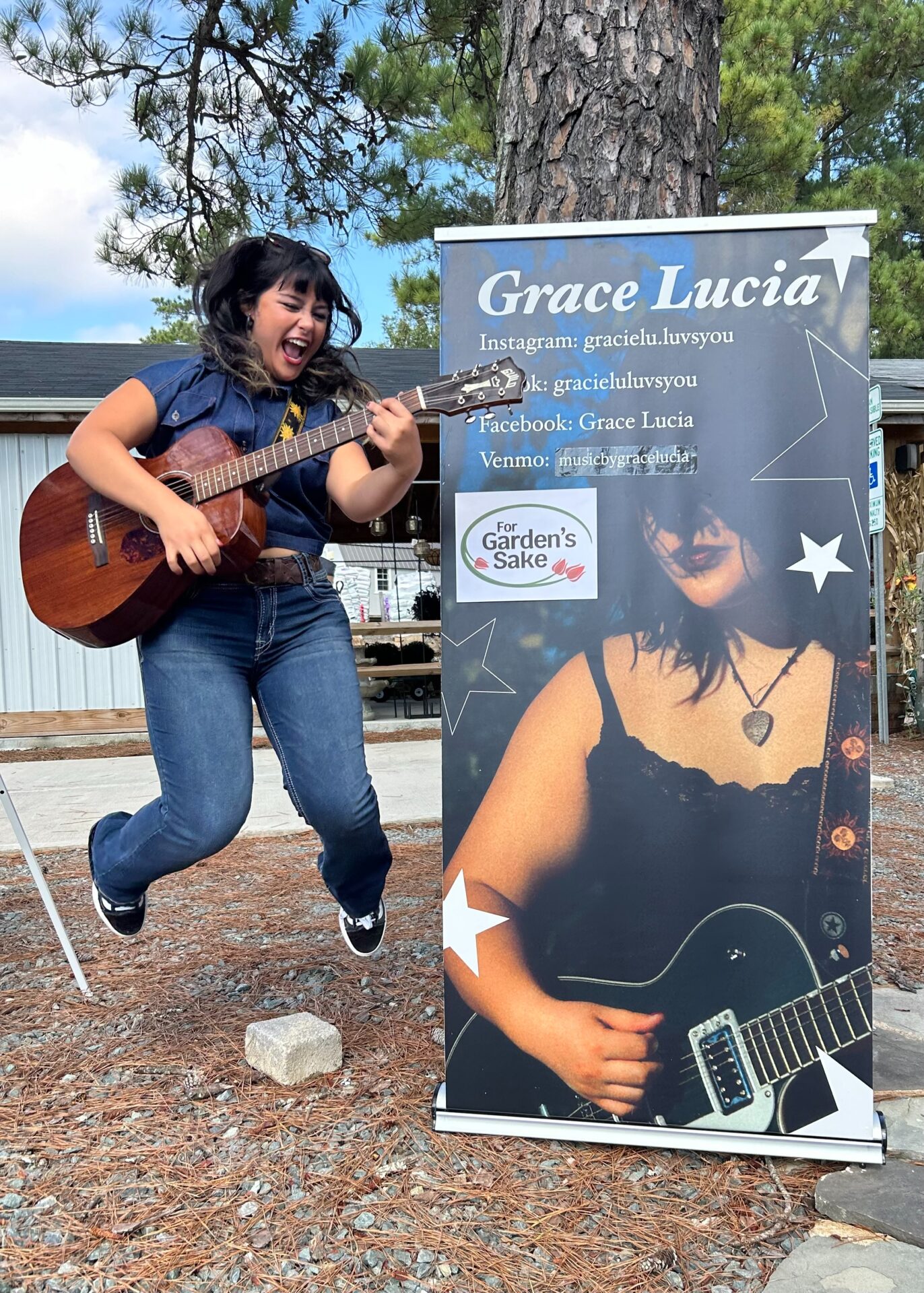 A person joyfully plays guitar near a promotional banner displaying social media information, outside a rustic building with trees in the background.