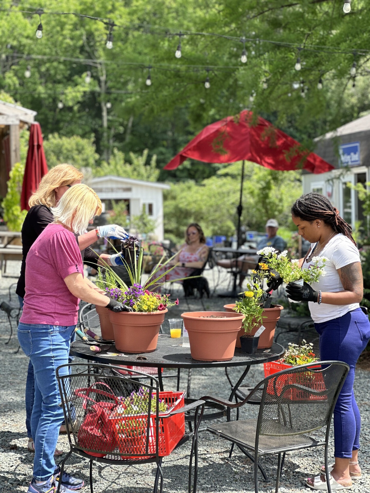 Three people plant flowers together outdoors at a garden center, with red umbrellas and greenery in the background. Tables and chairs are nearby.