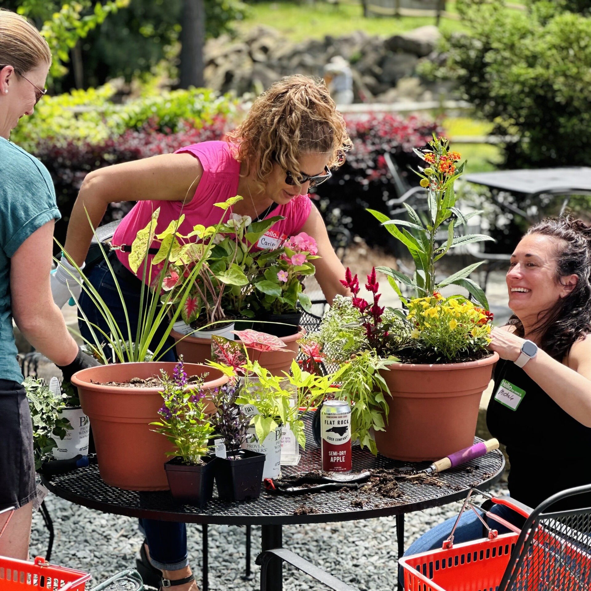 Three people are gardening together, arranging various plants in large pots outdoors. They are smiling and enjoying the sunny weather.