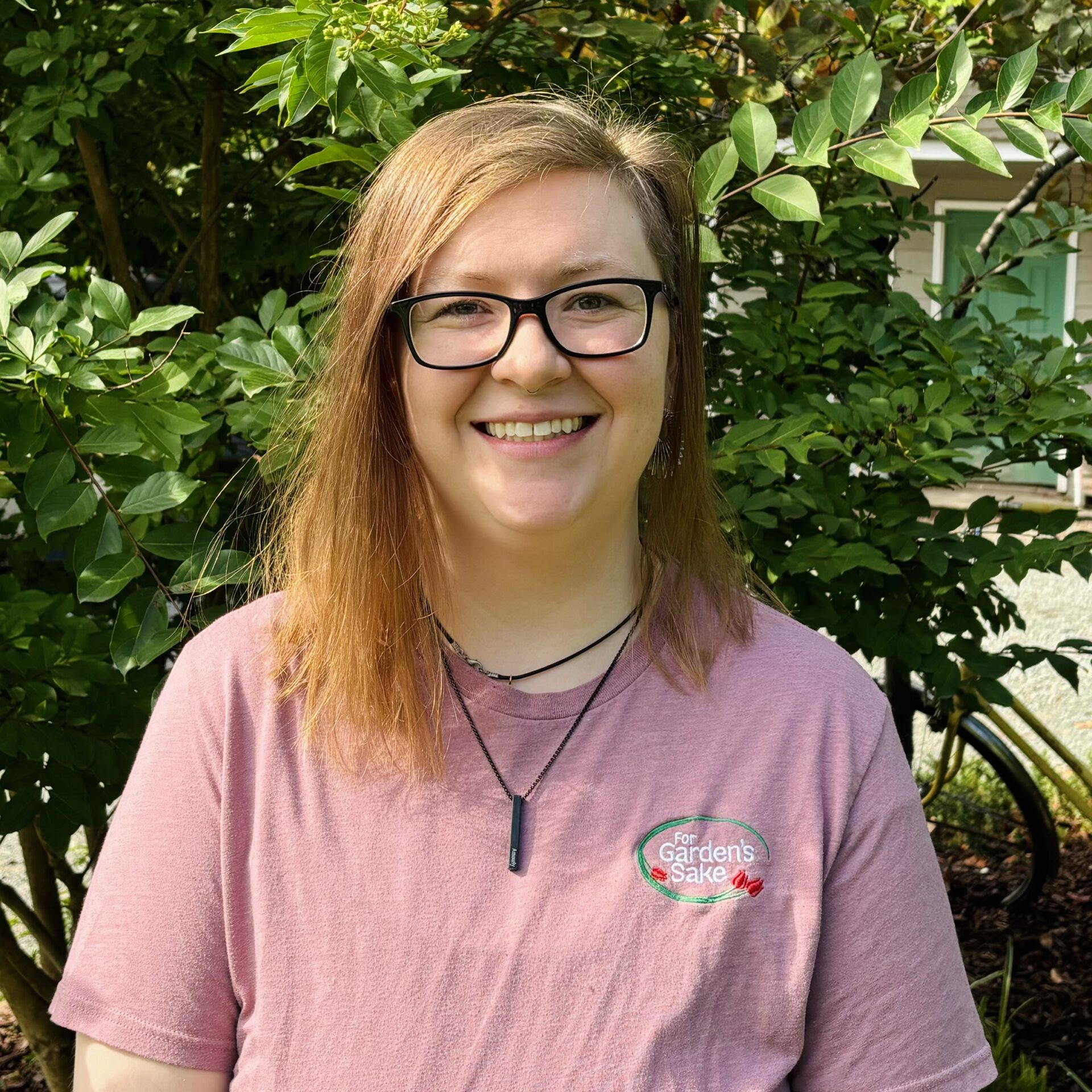 A person wearing glasses and a pink shirt stands in front of lush green foliage, smiling broadly with a building in the background.