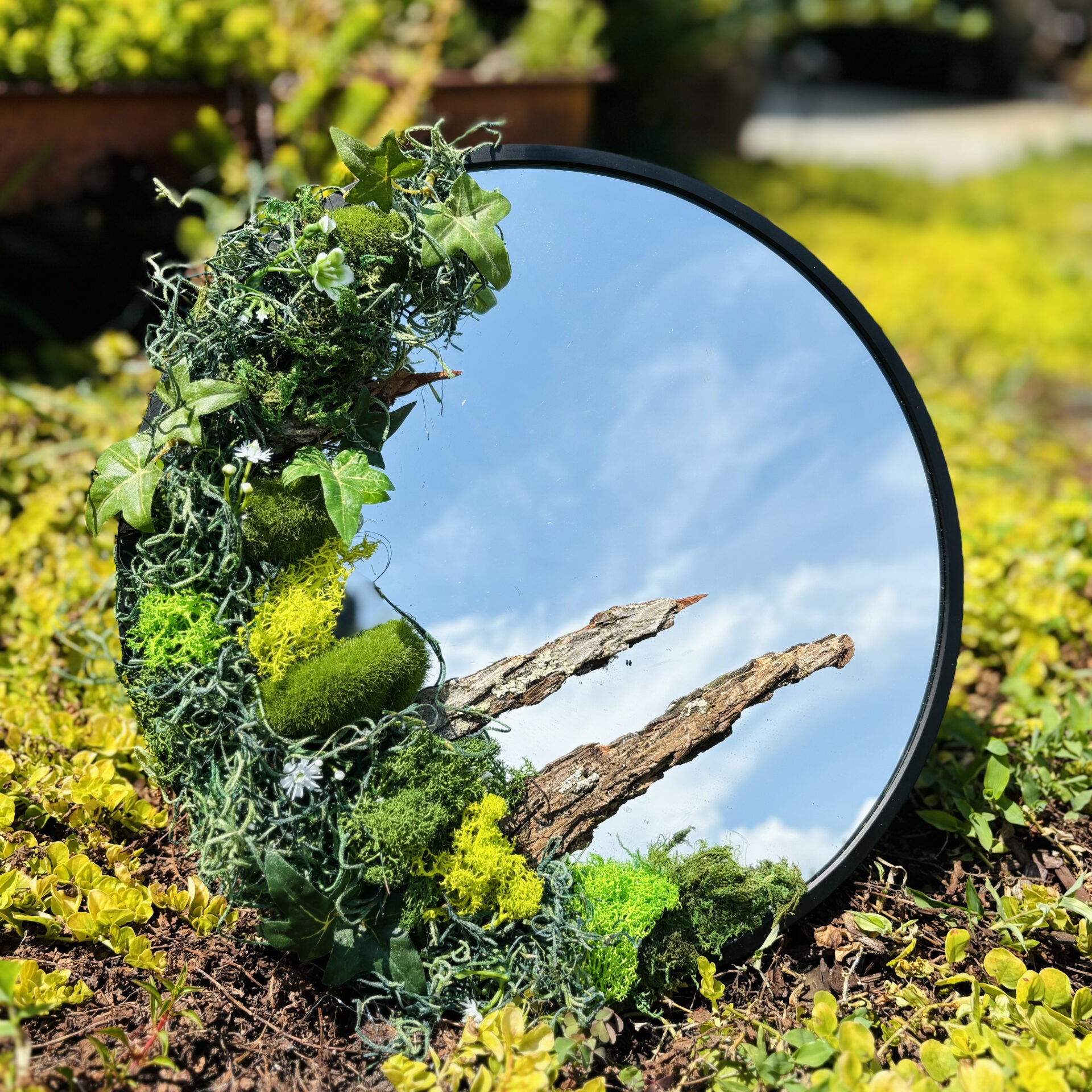 A round mirror decorated with greenery reflects the sky and branches, placed on the grass amidst a sunlit garden.