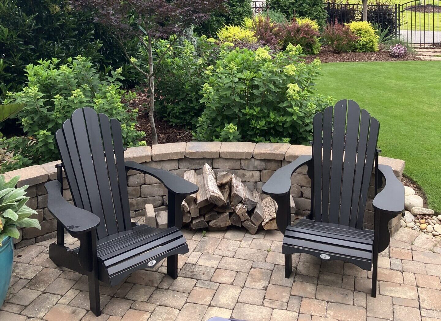 Two black Adirondack chairs on a stone patio, surrounded by lush greenery and a small stone wall. Stacked firewood is placed nearby.