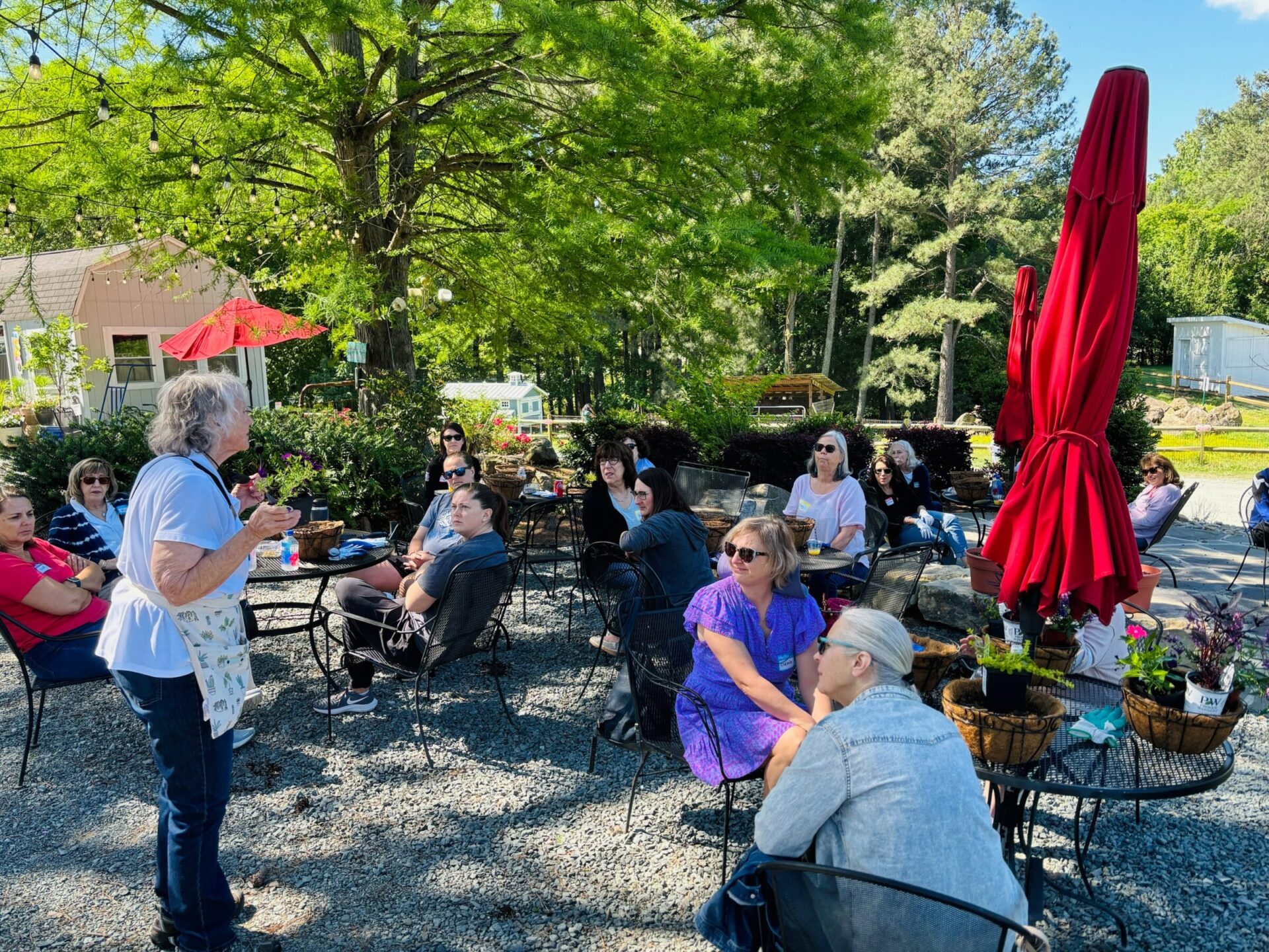 A group of people sitting outdoors near red umbrellas, listening to a person speak. Trees and greenery surround the casual garden setting.