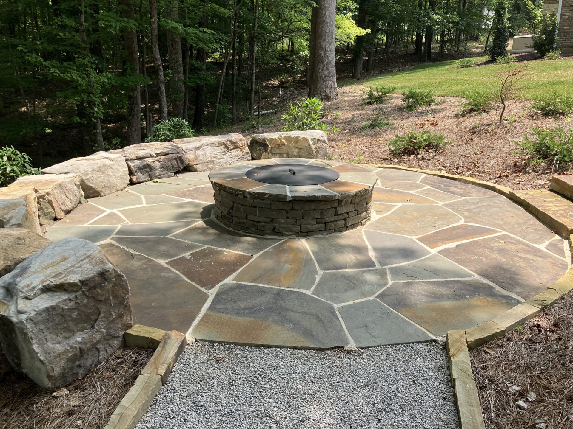 A circular stone patio with a central fire pit surrounded by greenery and trees, featuring large rocks on its perimeter.