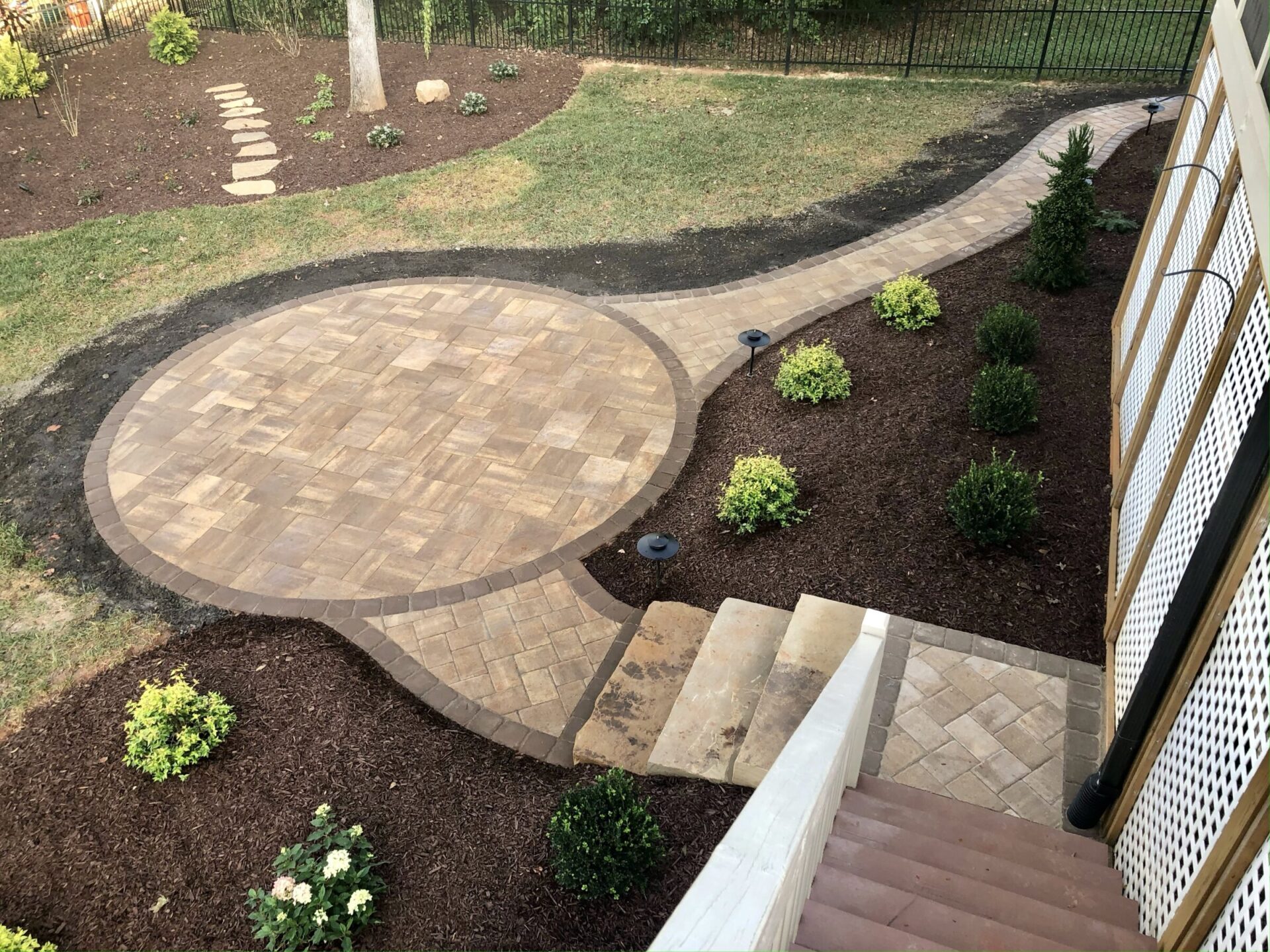 Brick patio with a circular design, surrounded by landscaped garden beds. Stone pathway and steps lead to the area. Green shrubs and mulch present.