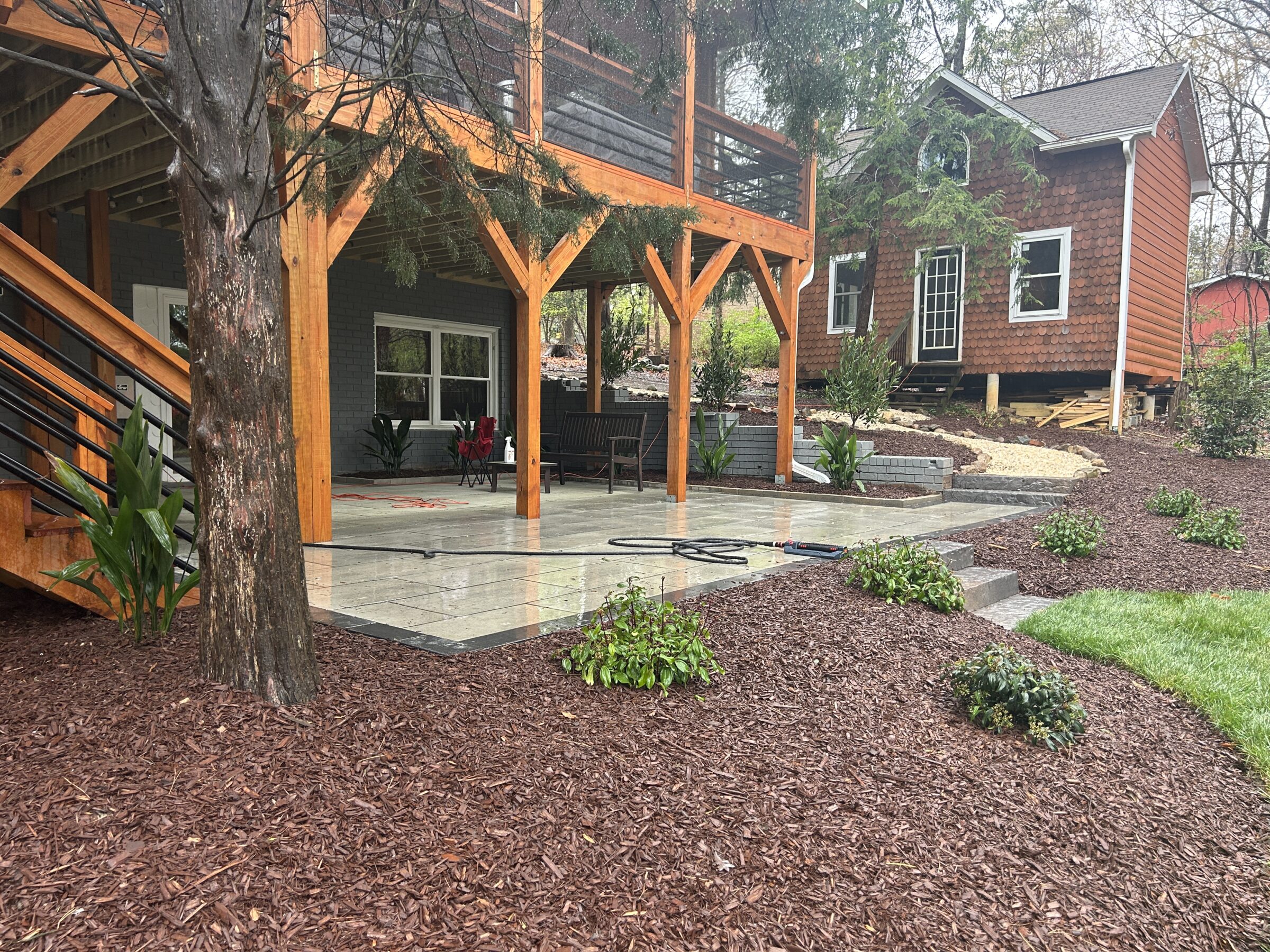 A porch with wooden beams overlooks a landscaped yard. A small building is visible in the background. No recognizable landmarks or historical buildings.