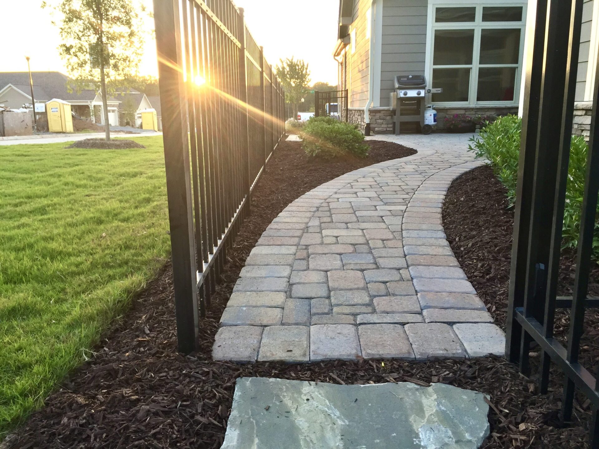 A paved stone pathway curves alongside a house with a barbecue grill. The sun sets, casting warm light on the manicured lawn.